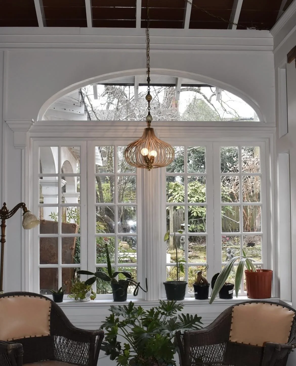 View of a sunroom or porch with large window and glass doors, several potted plants on the windowsill, a hanging light fixture, and chairs in the foreground.