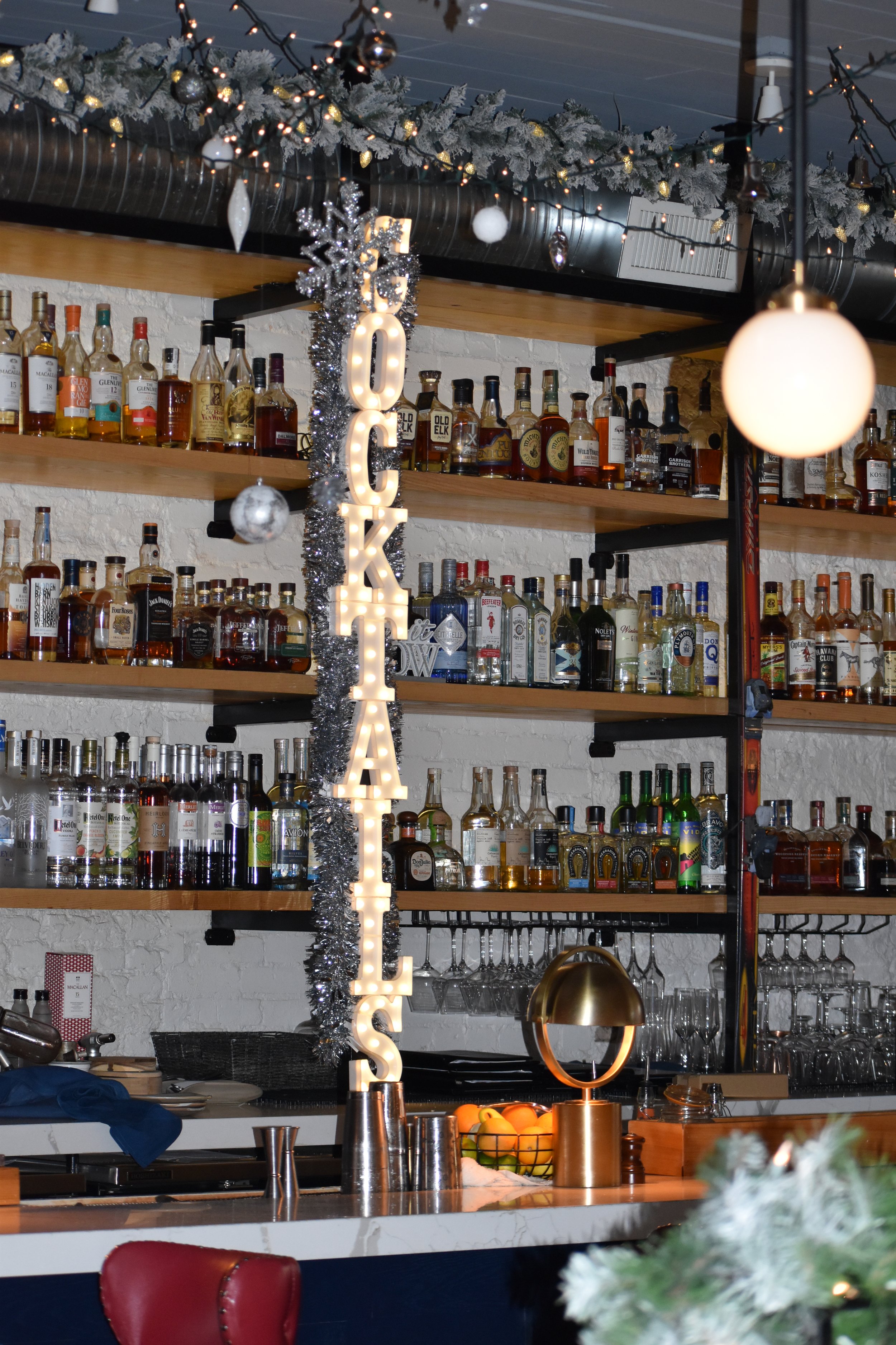 A bar decorated for Christmas with a lit sign spelling 'COOKIE' hanging vertically, surrounded by silver tinsel. Christmas garland and ornaments are hung along the ceiling. Shelves filled with various bottles of alcohol are visible behind the bar. A 