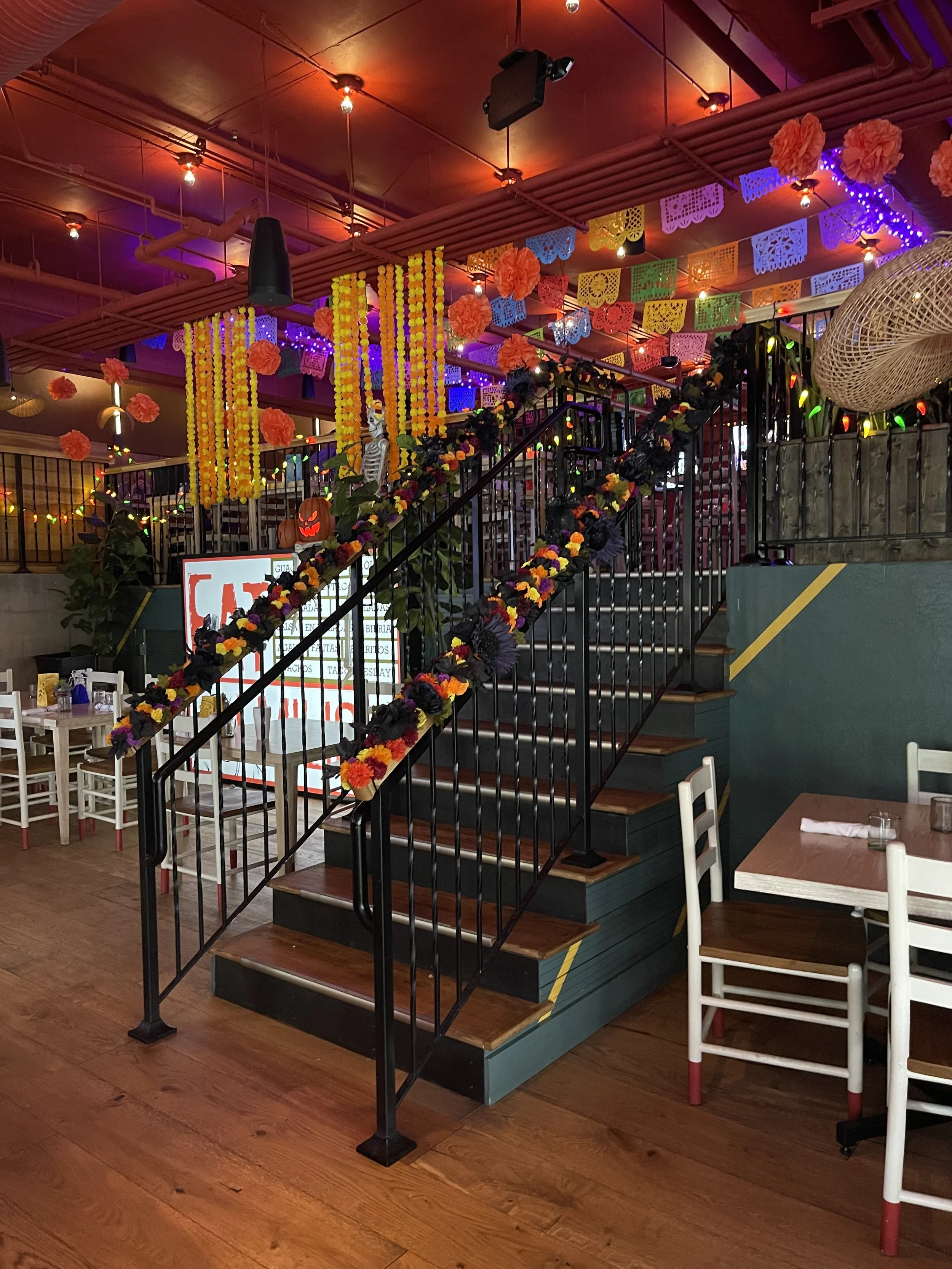 Decorative celebration area with vibrant hanging paper flowers, colorful paper banners, string lights, and floral garlands on a staircase in a lively restaurant or event space. Emilio's Tacos & Tequila Midtown Atlanta.