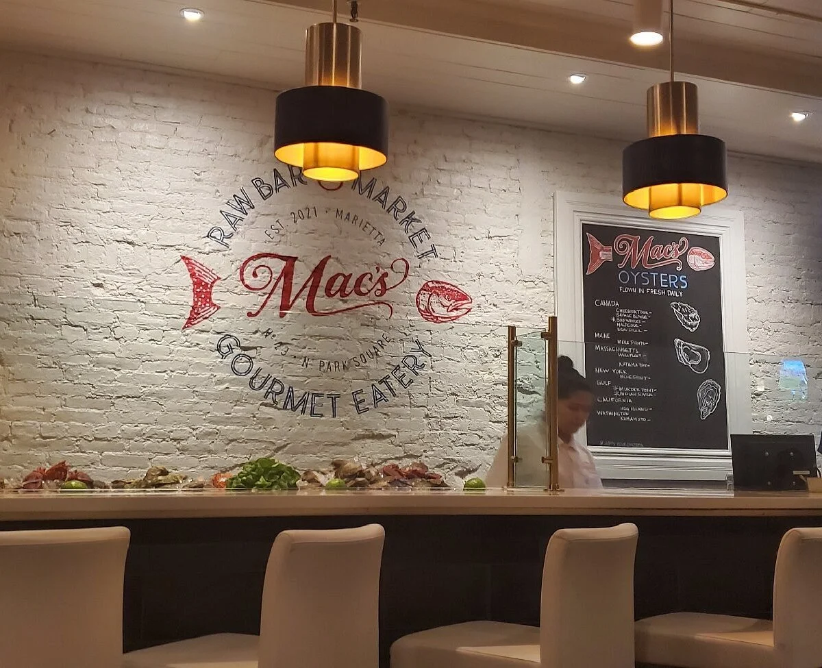 Interior of Mac's seafood restaurant with a white brick wall, illuminated pendant lights, a chalkboard menu, and a bar counter with seafood displayed, in Marietta, Georgia.
