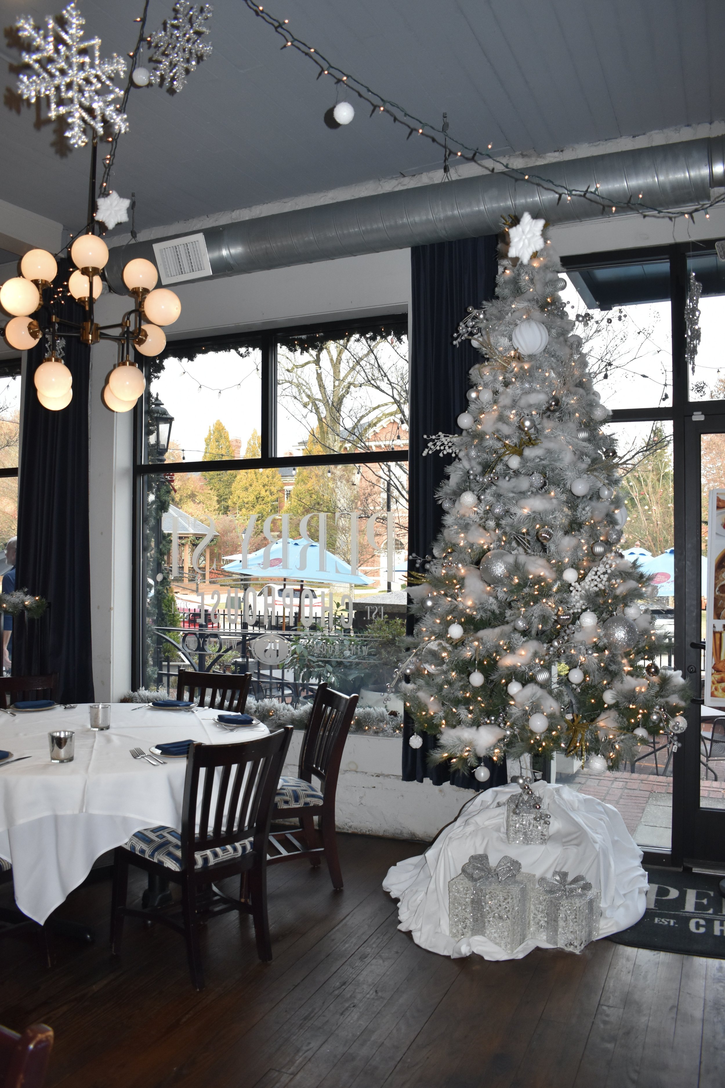 Decorated white Christmas tree with ornaments and lights inside a restaurant near large windows, with dining tables and chairs. Perry St Chophouse.