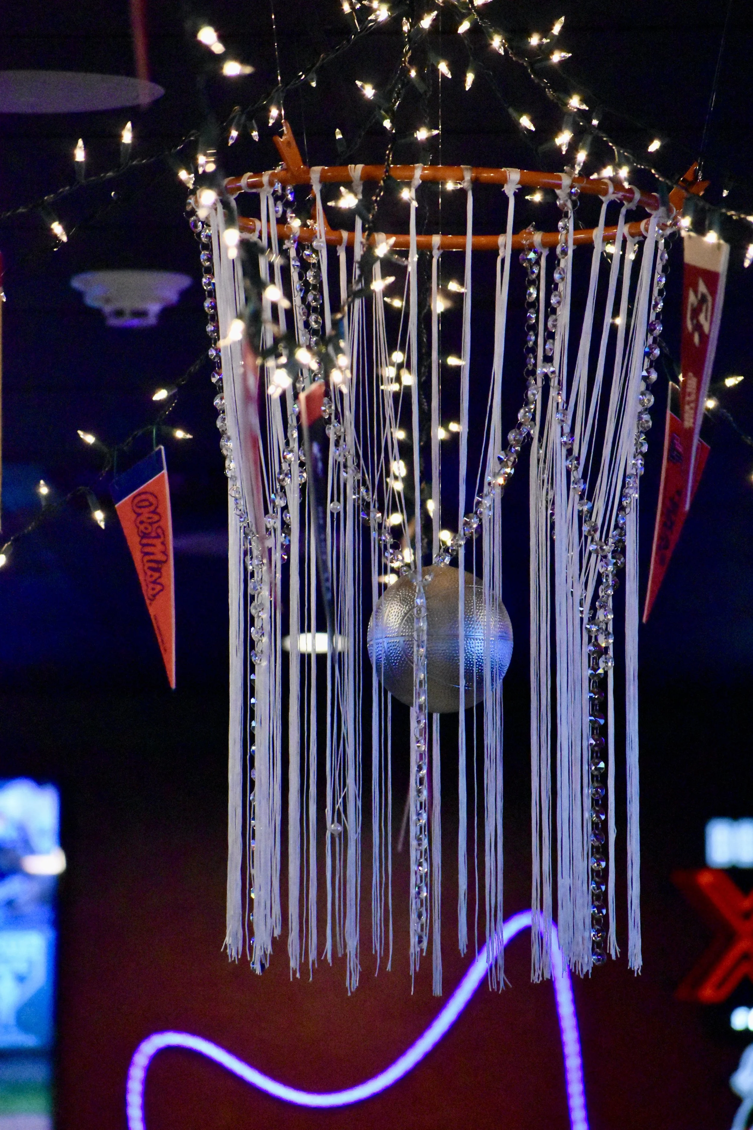 A decorative chandelier with strings of beads, a disco ball, and string lights hanging from a dark ceiling. March Madness