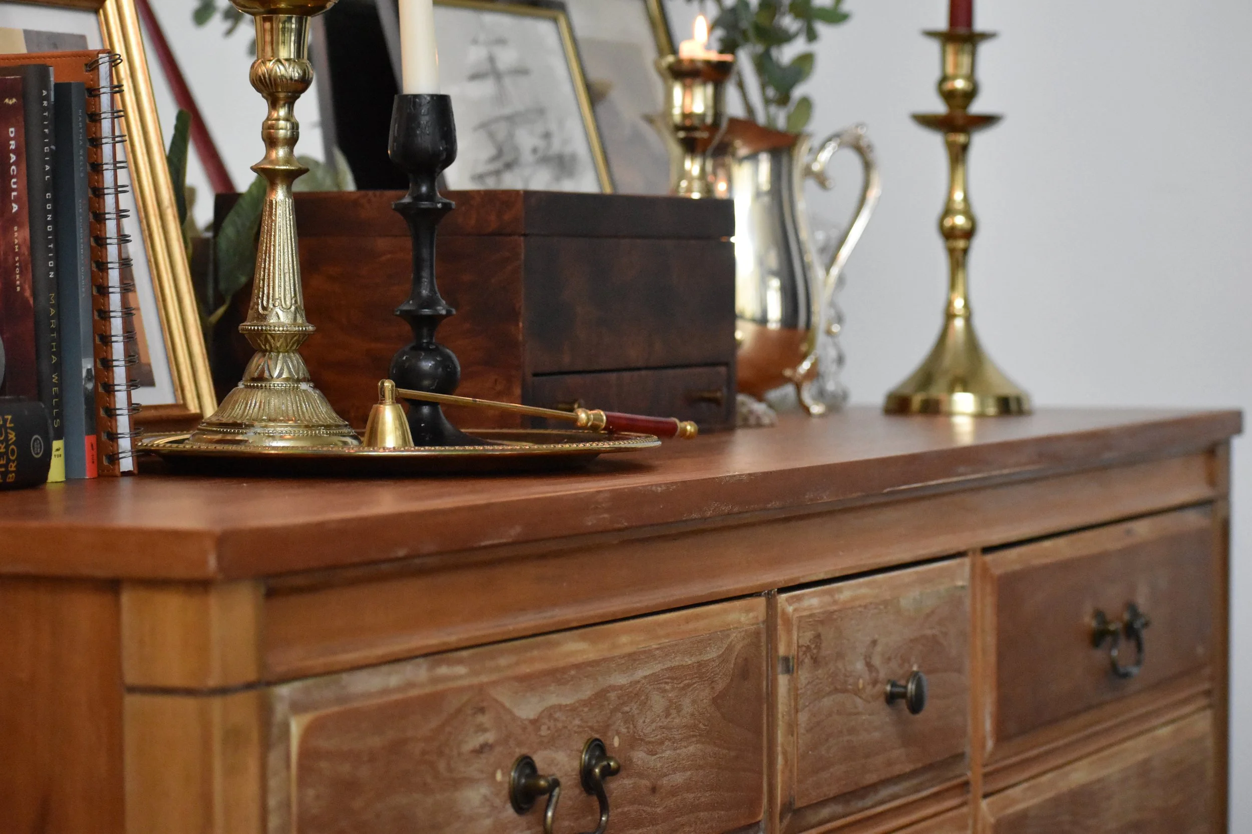 A wooden sideboard decorated with vintage items including brass candlesticks, a black candlestick, a tray, a wooden box, books, and a framed sketch, with a mirror and a plant in the background.
