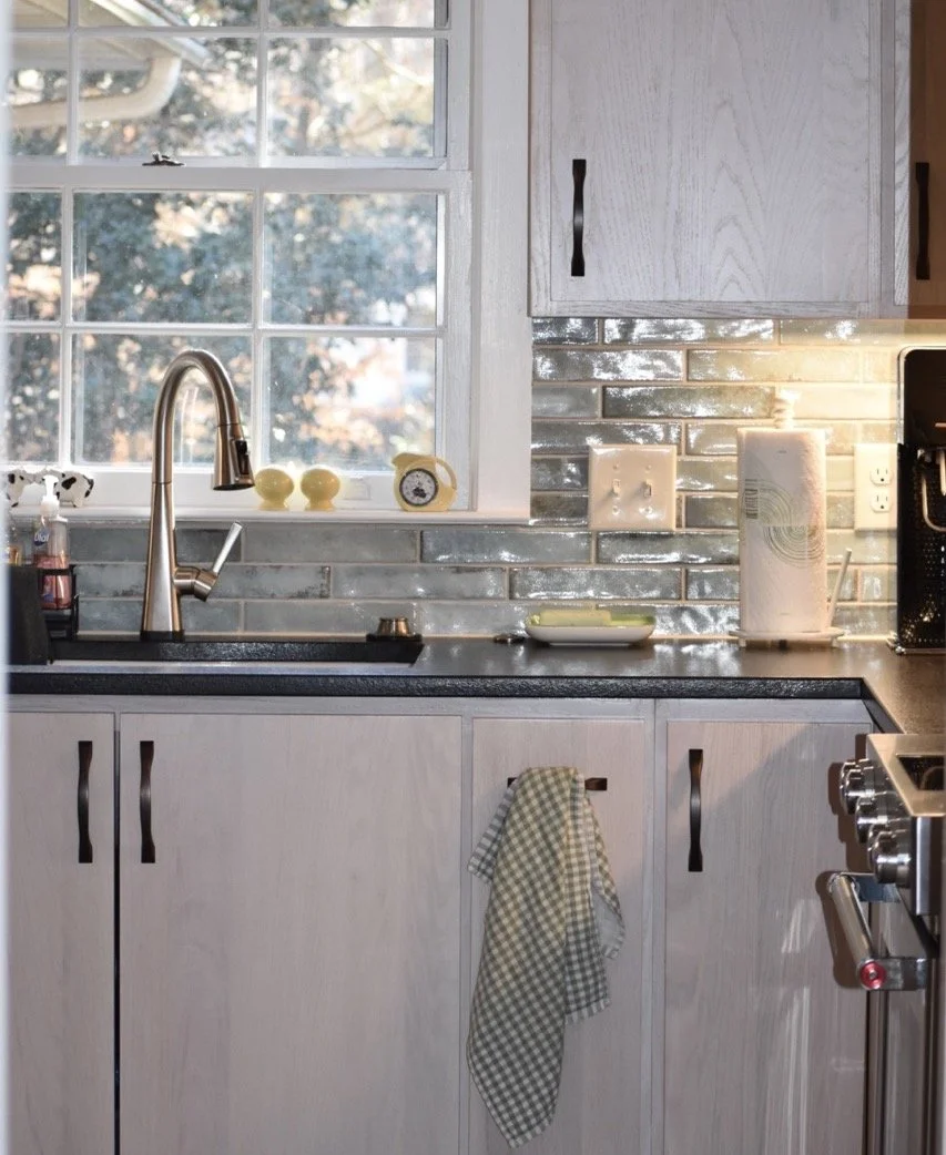 A kitchen window with a view of trees outside, a kitchen sink with a silver faucet, a small clock, and two yellow decorative objects on the windowsill. There are light-colored cabinets, a black countertop, and a backsplash with shiny gray tiles. A pa