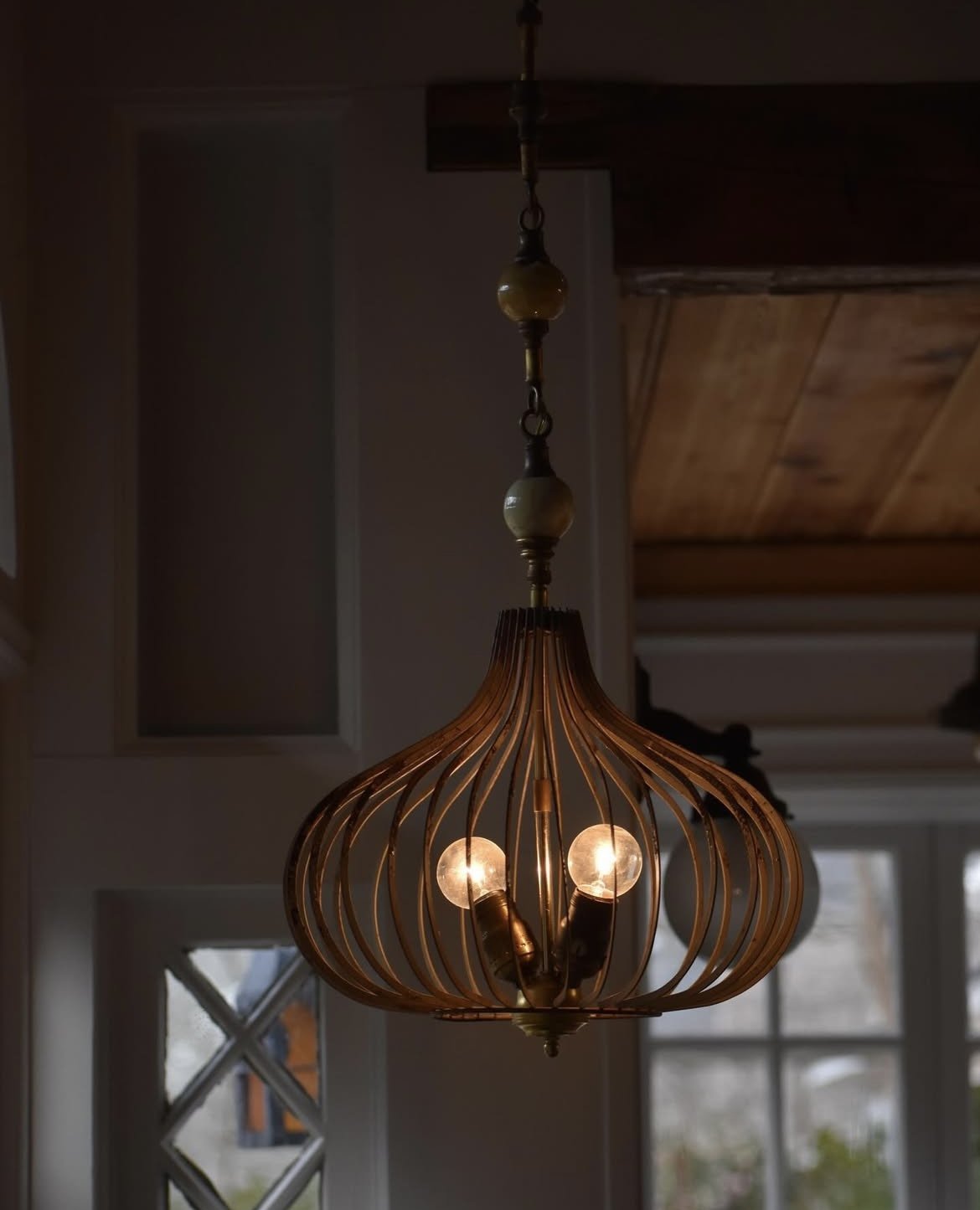 A vintage hanging chandelier with a wooden curved frame, holding three exposed light bulbs, in a room with large windows and a wooden ceiling.