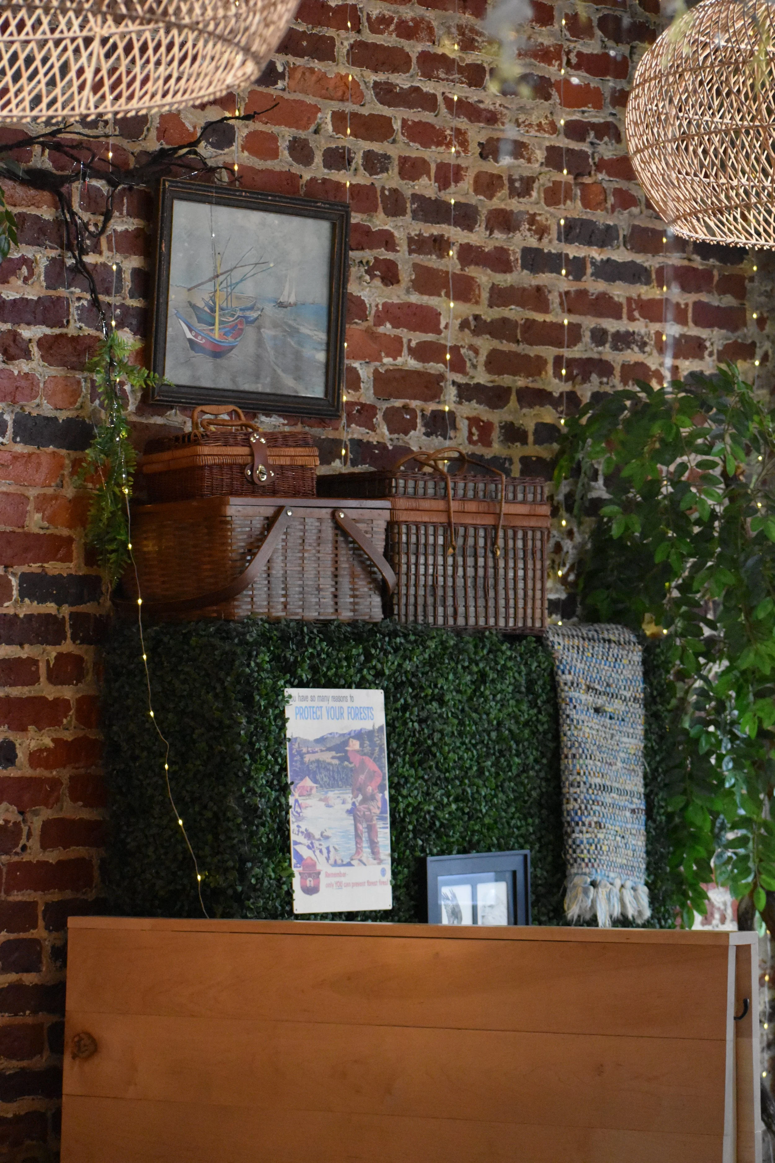 Decorative indoor scene with a brick wall, framed painting of boats, wicker baskets, fairy lights, and green plants. Hamp & Harry's Marietta Square.