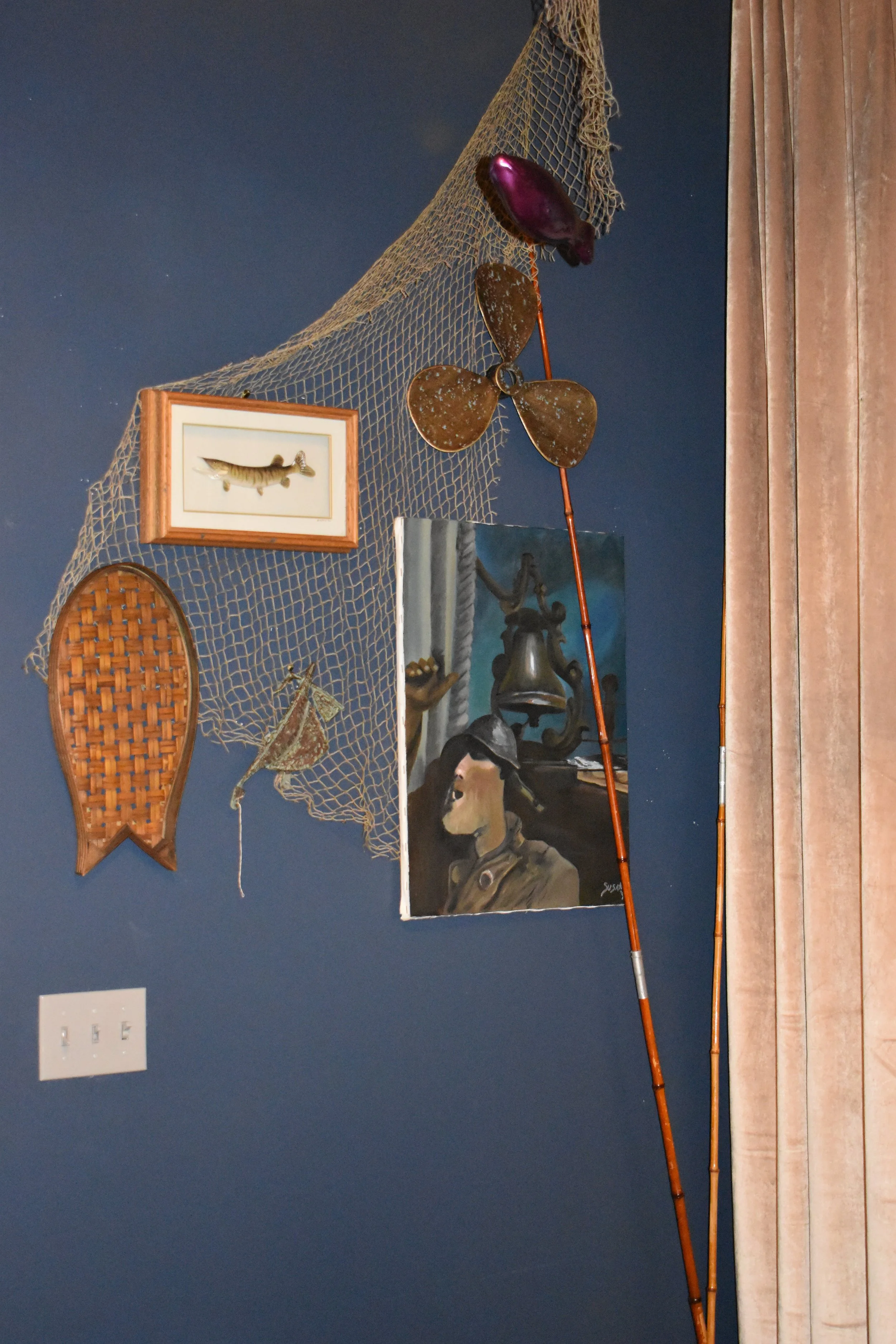 Decorative wall arrangement with fishing-themed items, including a fishing net, a framed fish illustration, a small woven basket, a mounted moth, a painted dog portrait, bamboo fishing rods, and a curtain on a blue wall. Hamp & Harry's Marietta