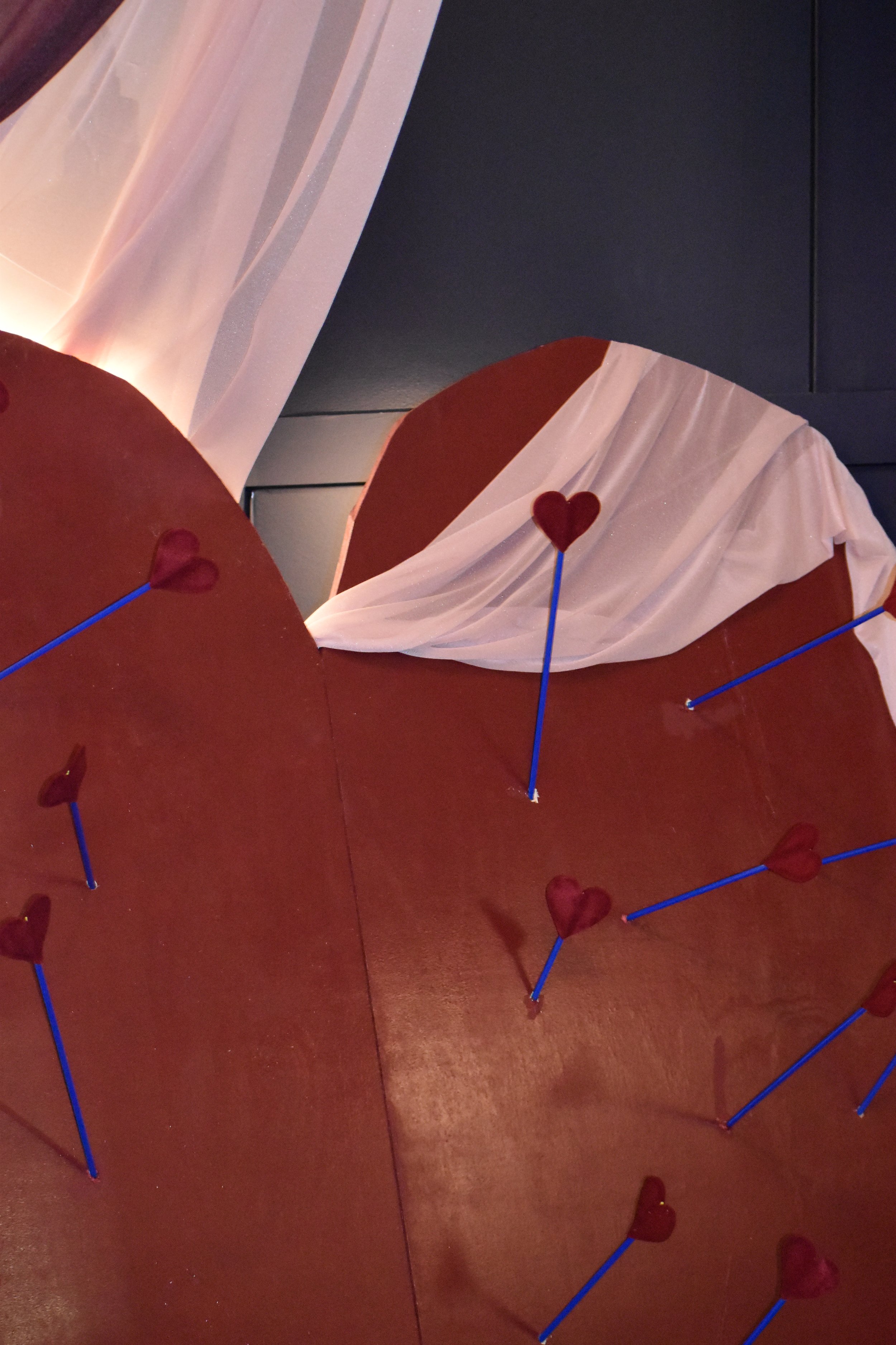 Close-up of a large red heart-shaped object decorated with multiple small red heart-shaped pins on blue sticks, with a white sheer fabric draped in the background.