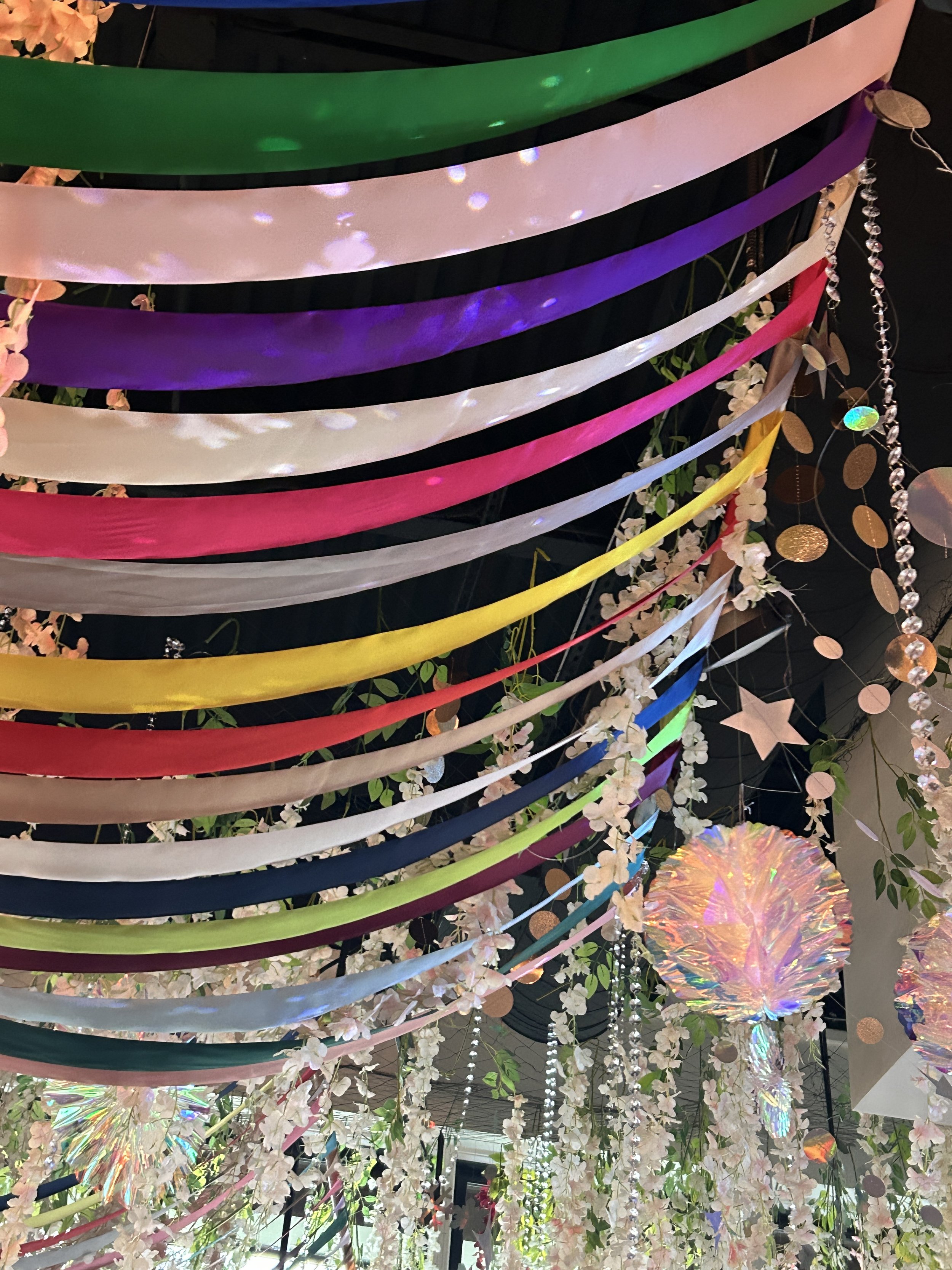 Colorful hanging ribbons and decorations, including flowers, stars, and iridescent spheres, create a festive ceiling decoration.