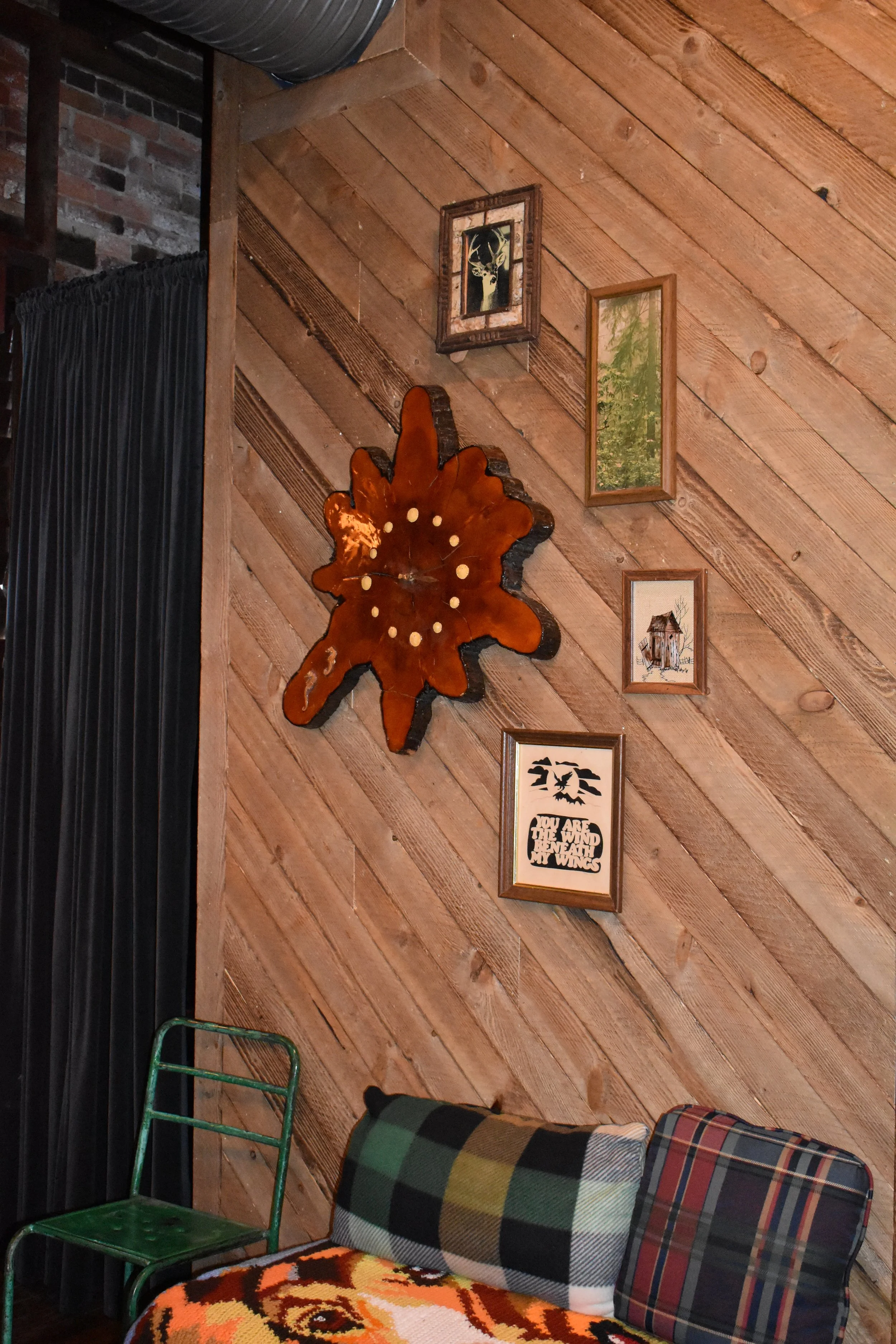 Wood-paneled wall decorated with framed pictures and artwork, including a wooden clock shaped like a maple leaf, with a green chair, a plaid and checkered cushion, and a blanket with a tiger pattern in the foreground.