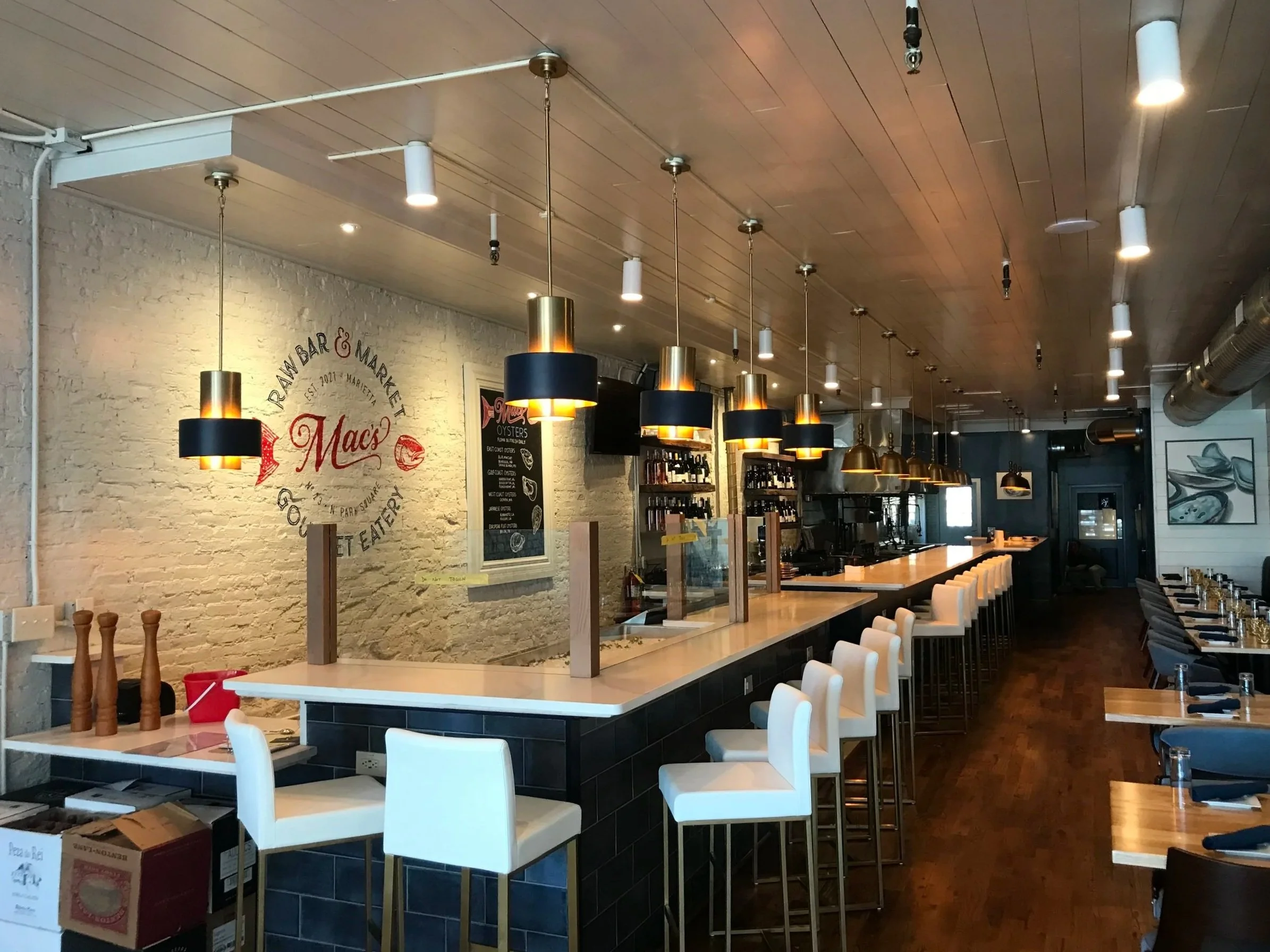 Interior view of a restaurant with a long bar, white chairs, and black hanging lamps with gold accents, white brick walls, and wooden ceiling.