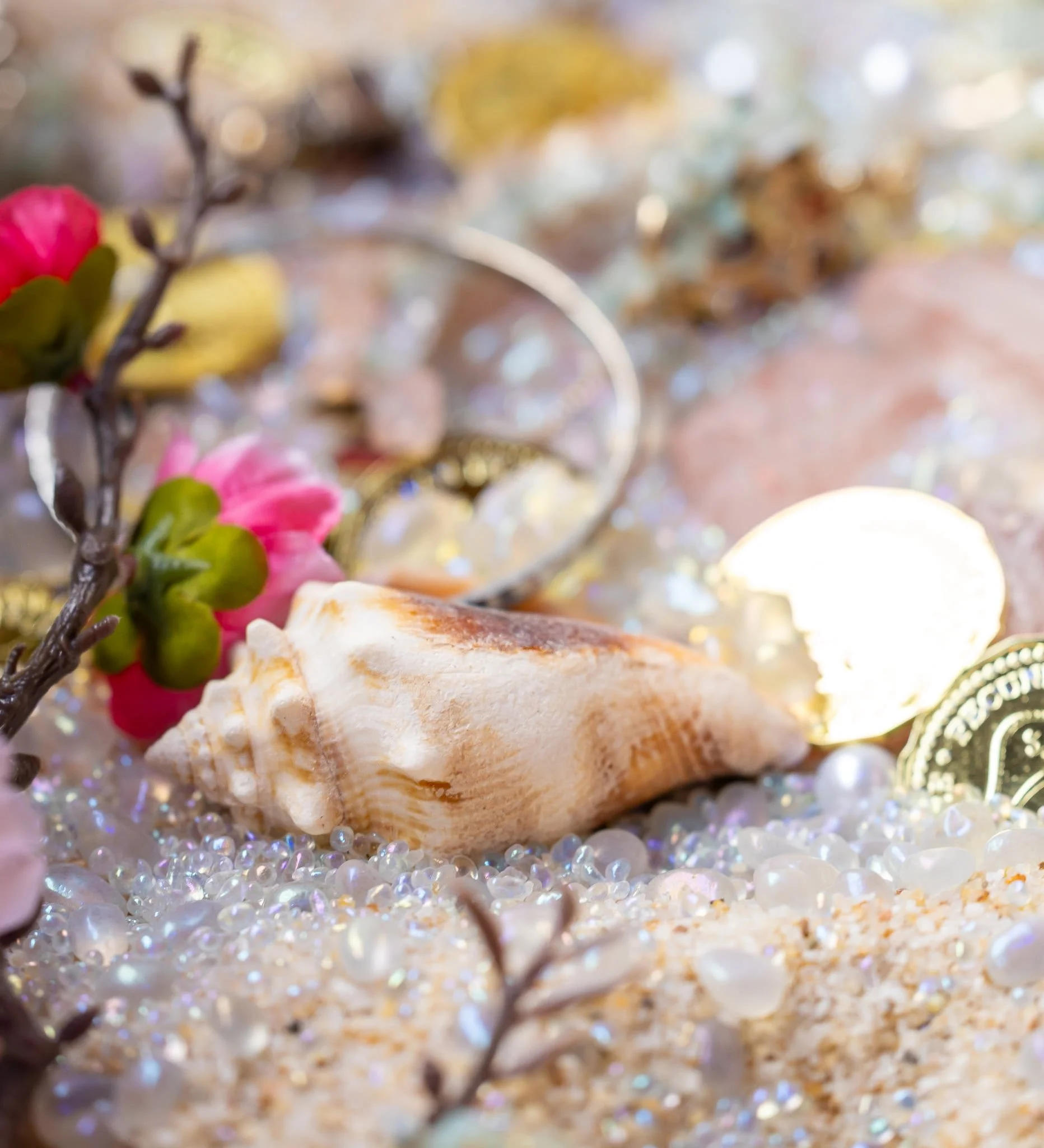Close-up of seashells, pearls, rocks, and small pink flowers on a sandy surface with sparkling elements.