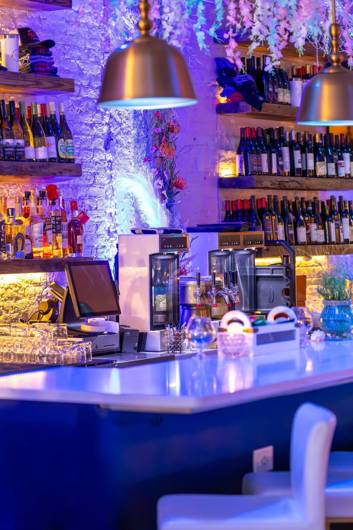 Interior of a bar with bottles of alcohol on shelves, a white bar counter with glasses, a computer, and various bar equipment; illuminated with colorful lighting and decorative flowers, hanging pendant lights overhead.