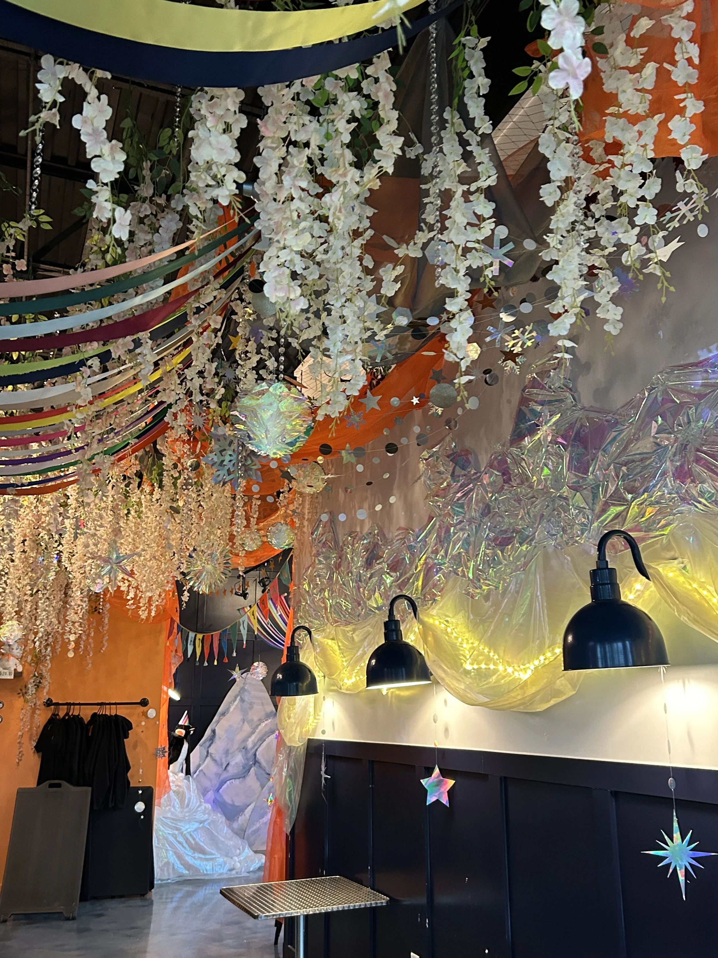 Decorated indoor space with hanging artificial white flowers, colorful paper streamers, iridescent foil decorations, and star-shaped hanging ornaments. There are black wall-mounted lights, a draped yellow fabric with string lights, and a mountain-lik