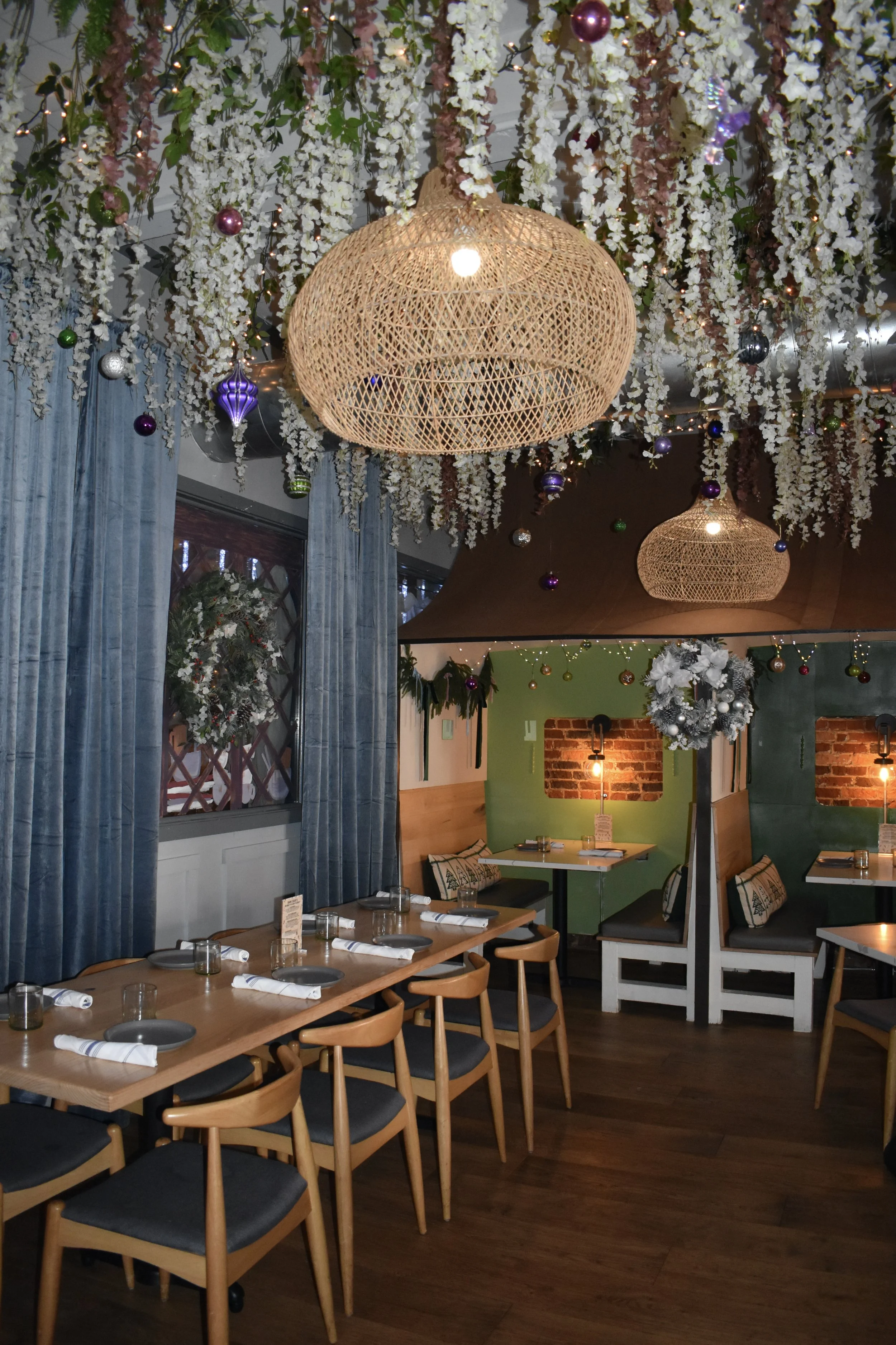 Decorated restaurant interior with hanging floral arrangements, woven pendant lights, Christmas ornaments, and set tables with napkins and glasses.