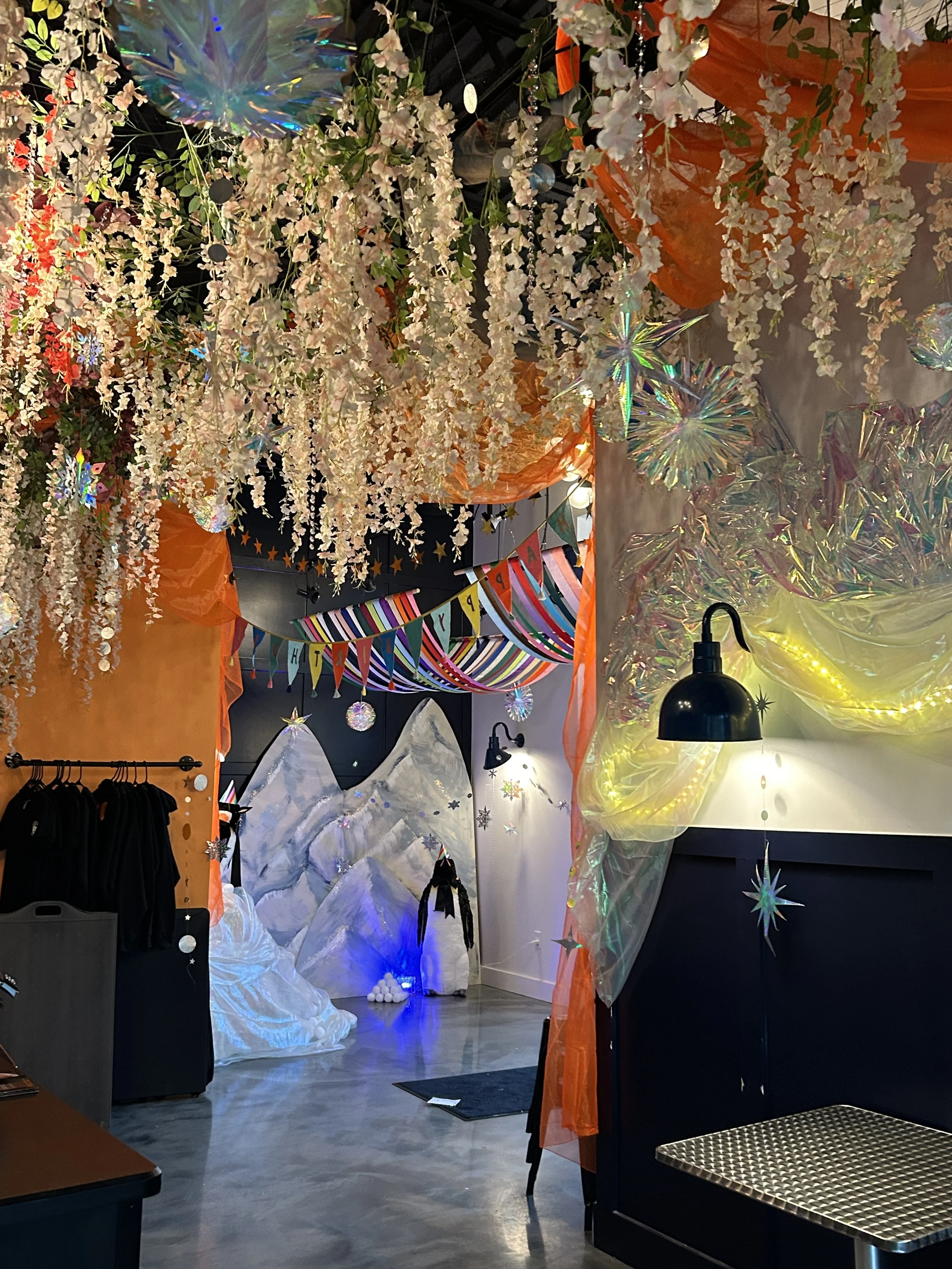 Colorful indoor space decorated with hanging white flowers and iridescent star-shaped ornaments, with a backdrop of snow-capped mountain art and rainbow-colored banners, illuminated by string lights and black wall lamps.