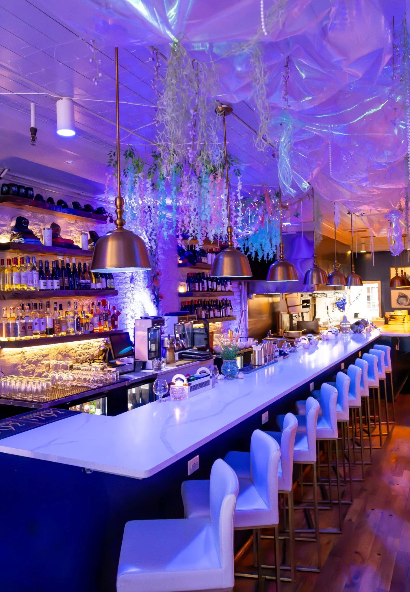 A modern bar with white countertops, white bar stools, and warm lighting. Shelves with various liquor bottles are mounted on the wall, with decorative hanging plants and string lights overhead, creating a cozy and stylish ambiance.