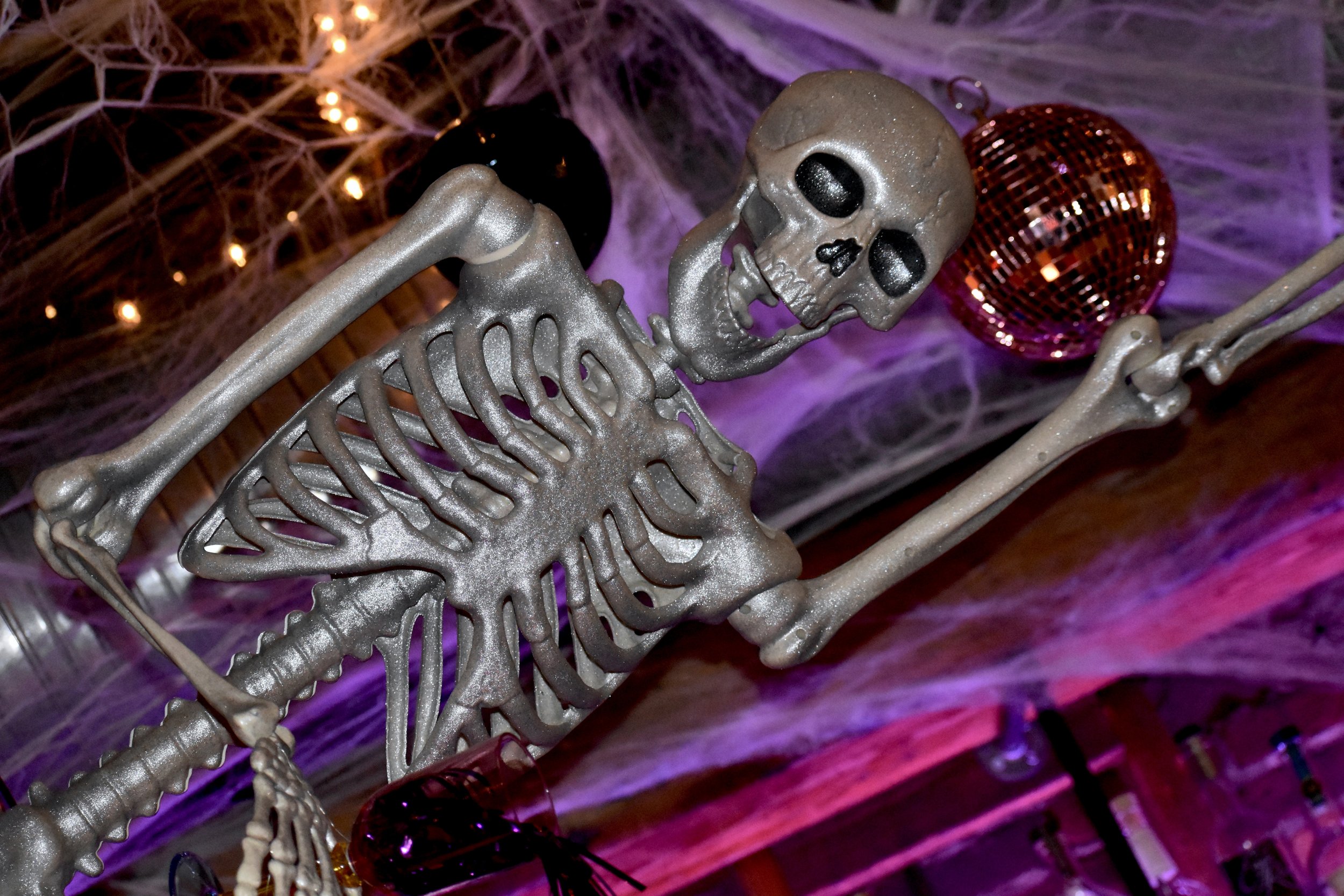 A Halloween decoration of a skeleton with silver paint, surrounded by purple spider web decorations and a red disco ball, illuminated by purple lighting.