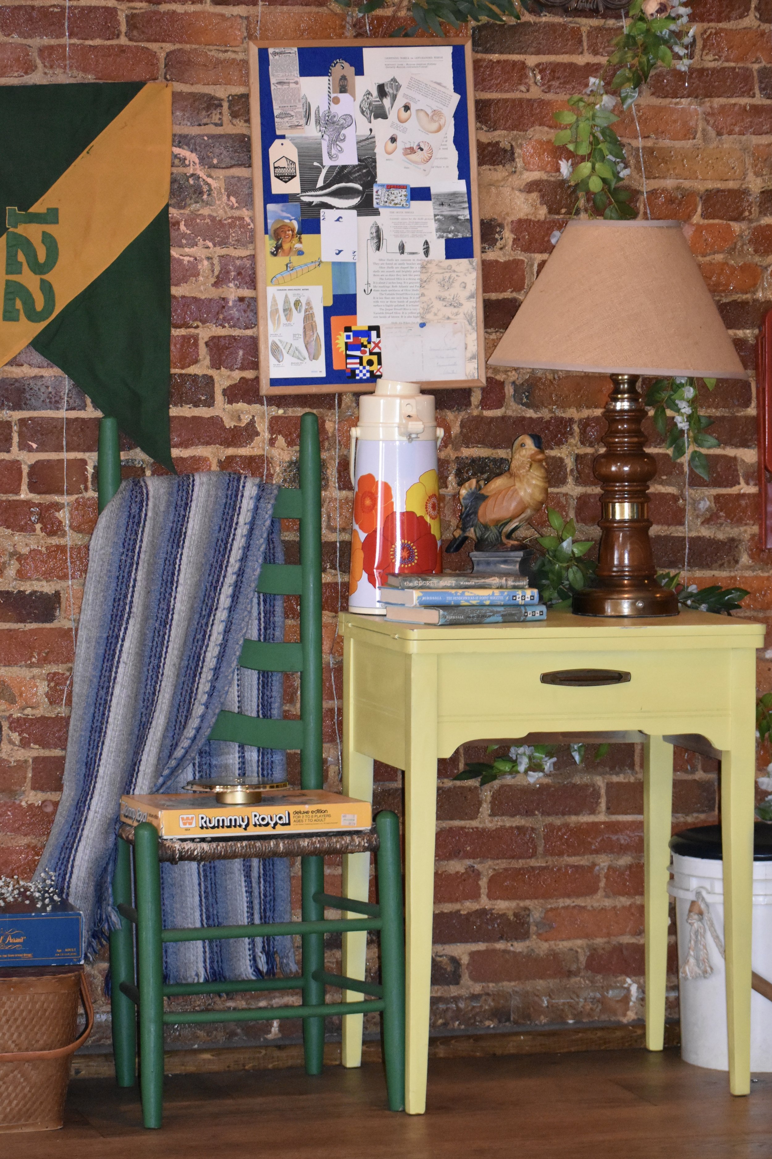 Interior decor scene with a brick wall, a green wooden chair with a blue striped cloth draped over it, a yellow table, a table lamp, a ceramic bird sculpture, a stack of books, a thermos with floral pattern, a cork bulletin board with various papers,