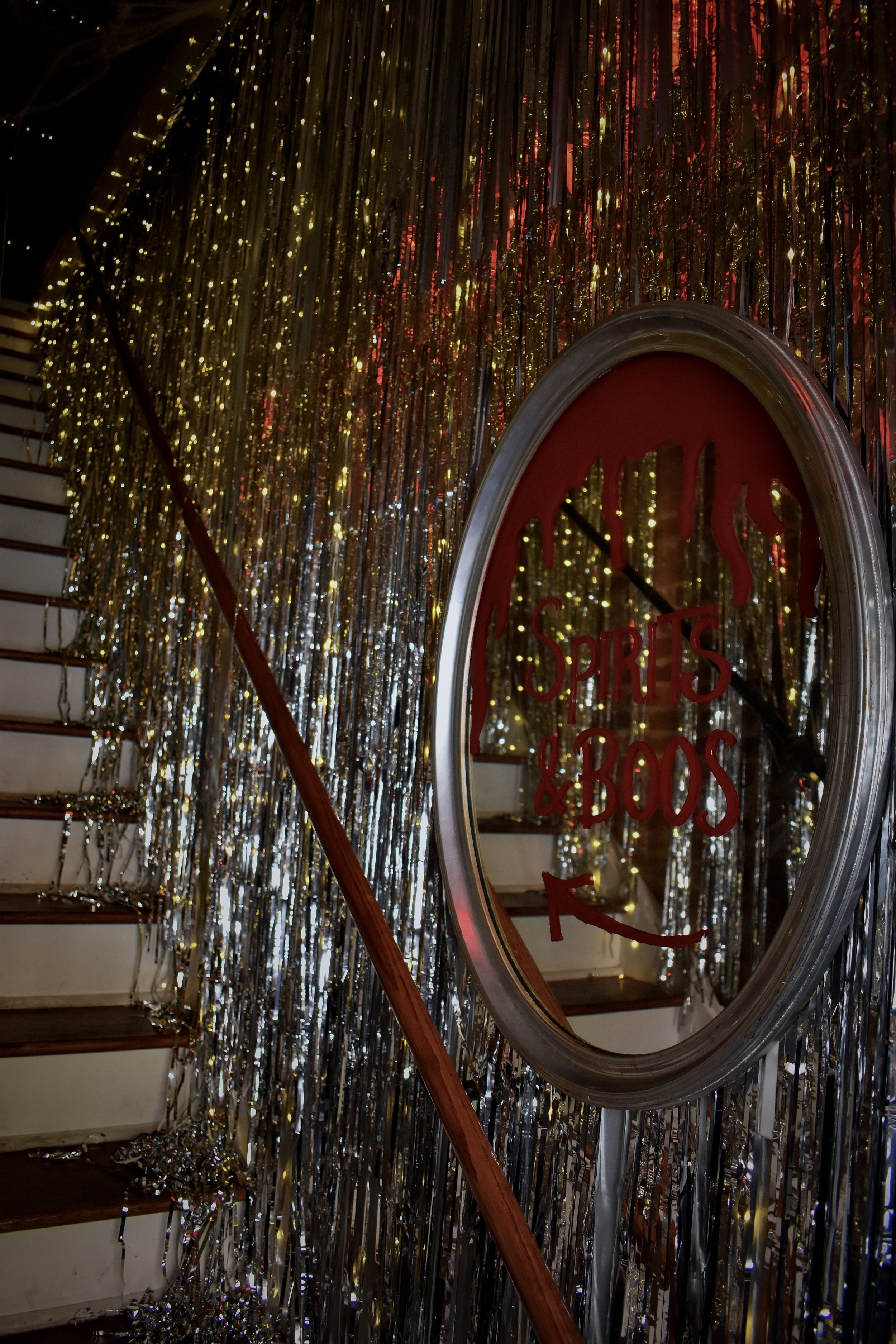 Decorated staircase with gold and silver streamers, a round mirror with red text that reads 'Spirits & Boos,' and a wooden handrail. Halloween decorations.