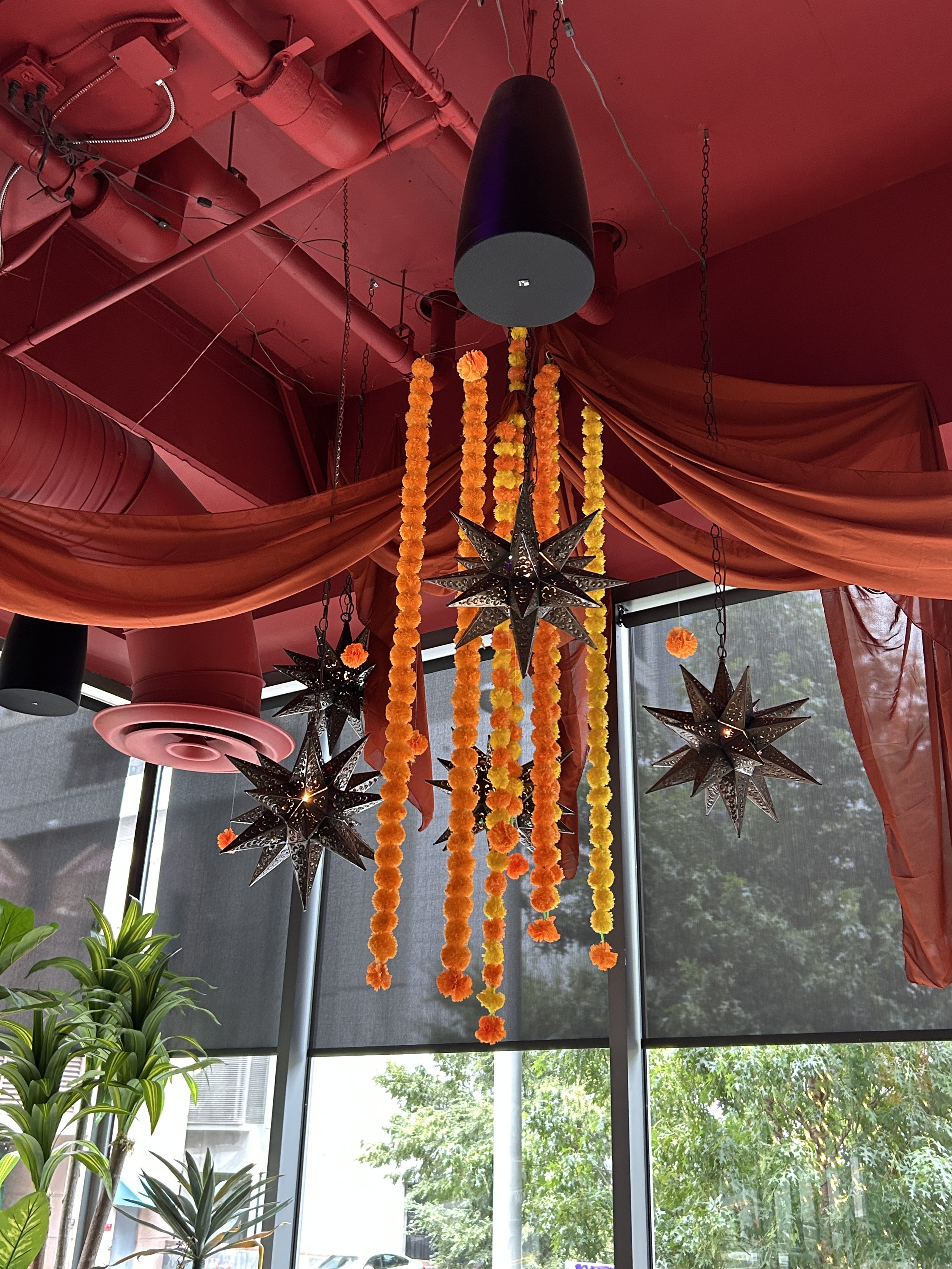 Decorative hanging star-shaped lanterns and orange and yellow marigold flower garlands hanging from the ceiling, with red fabric draped along the ceiling and large windows in the background showing trees outside.