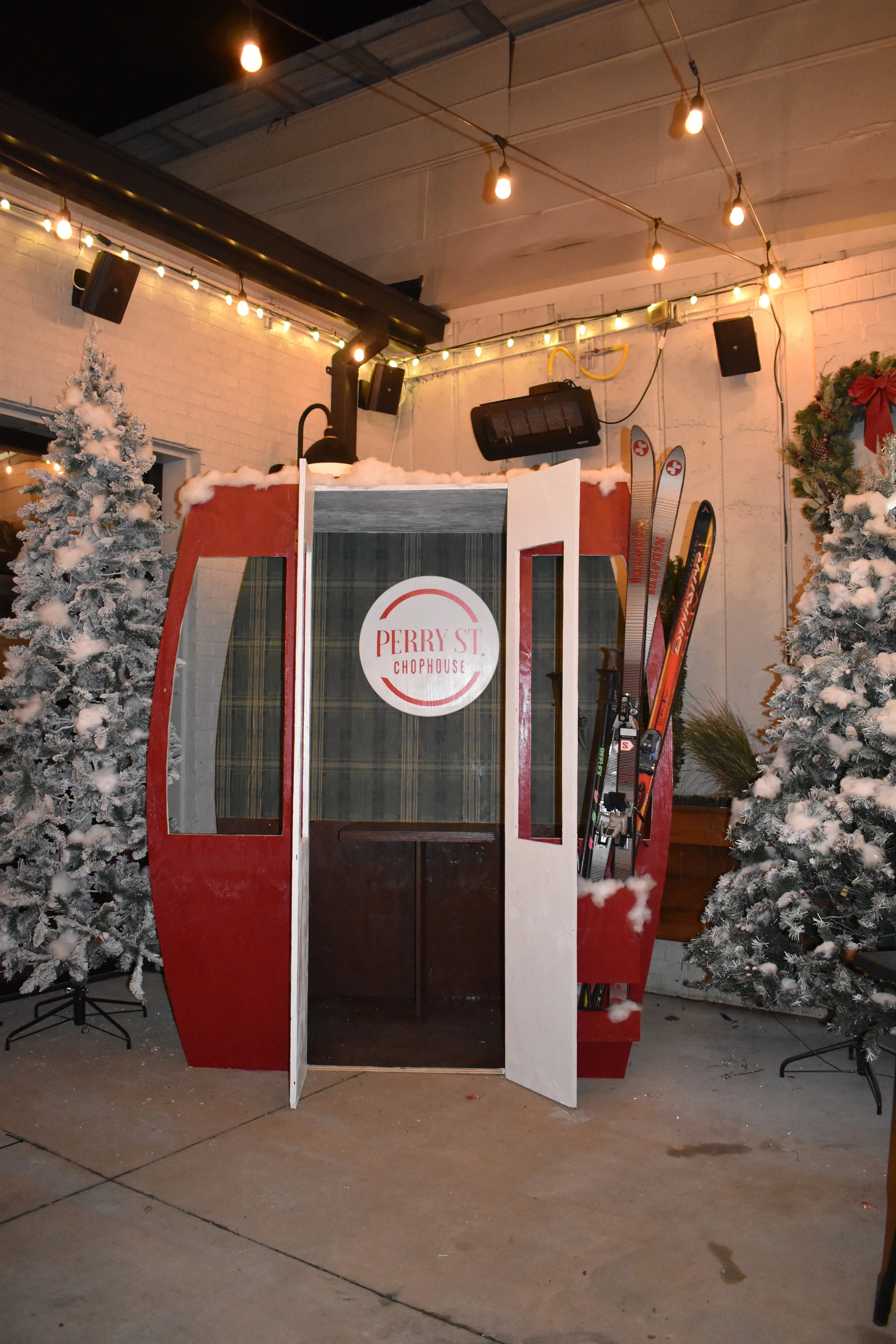 A red gondola-shaped photo booth with open doors, decorated with fake snow on the top, flanked by snow-covered Christmas trees, inside a decorated indoor space with string lights and skis mounted on the wall.