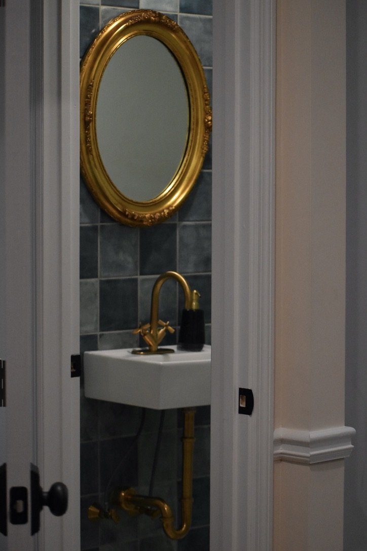 Small bathroom with a white sink, gold faucet, black soap dispenser, mirror with gold frame, gray tiled wall, and white molding around the doorway.