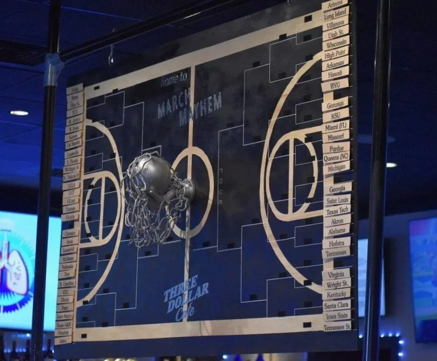 Basketball tournament bracket on a glass board with team names, featuring a small basketball hoop with a metallic basketball and net, illuminated by blue lighting.