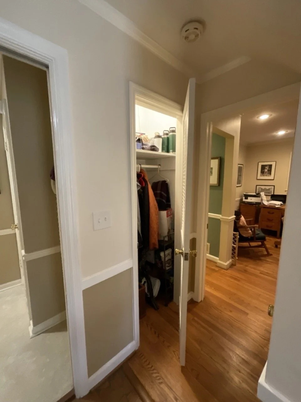 Open closet door revealing a small storage space with shelves, clothing, and containers. In the background, an office or study area with a desk, chair, and framed pictures on the wall.