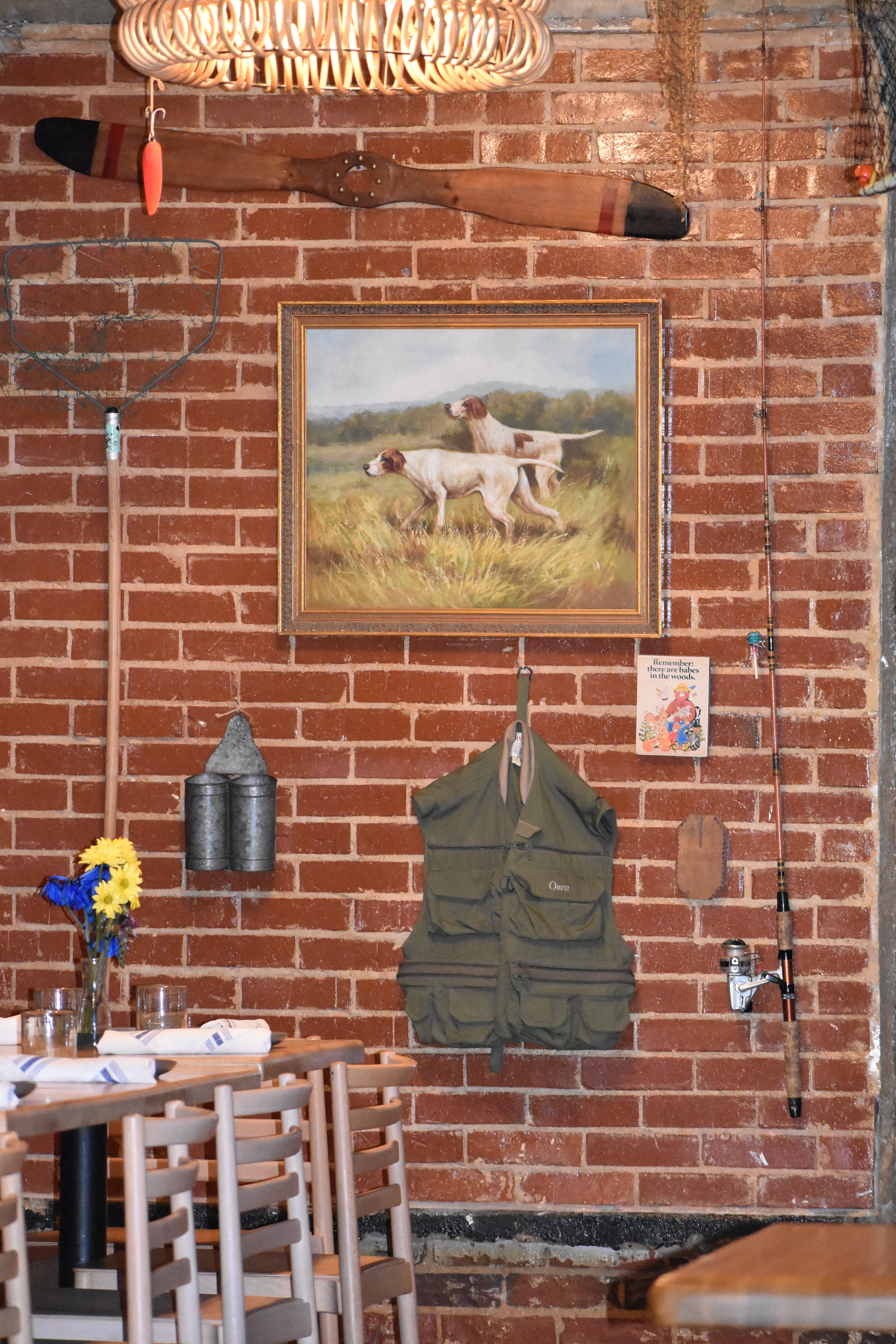 Decorative wall with a framed painting of two dogs running in a field, a vintage fishing rod, a fishing reel, a fishing tackle bag, a watering can with flowers, a weather vane, and a propeller, all mounted on a brick wall. Hamp & Harry's Marietta