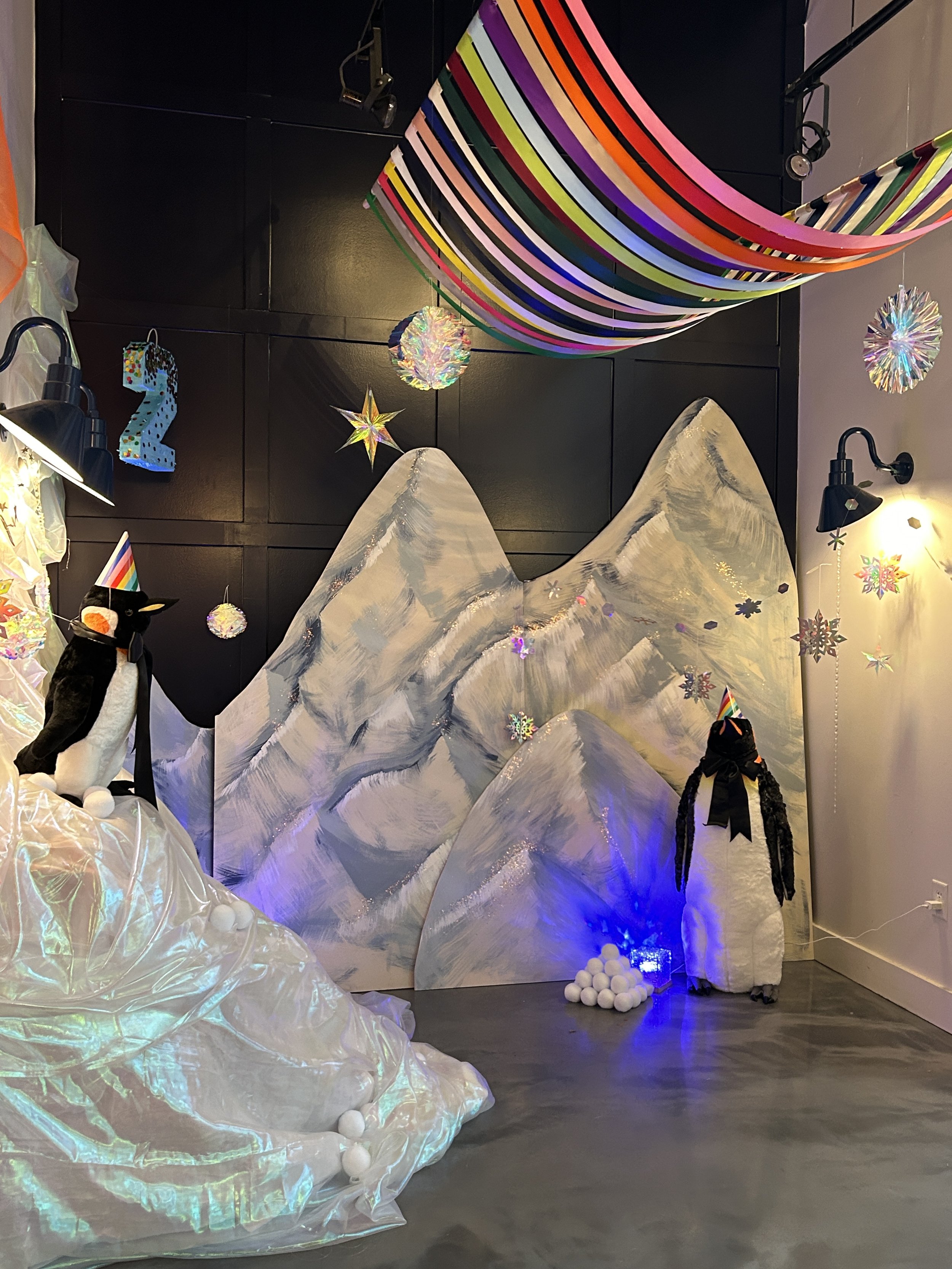 Indoor winter scene with painted mountain backdrop, two penguin decorations wearing party hats, a pile of white balls resembling snow, colorful hanging decorations, and rainbow-striped fabric draped from the ceiling.