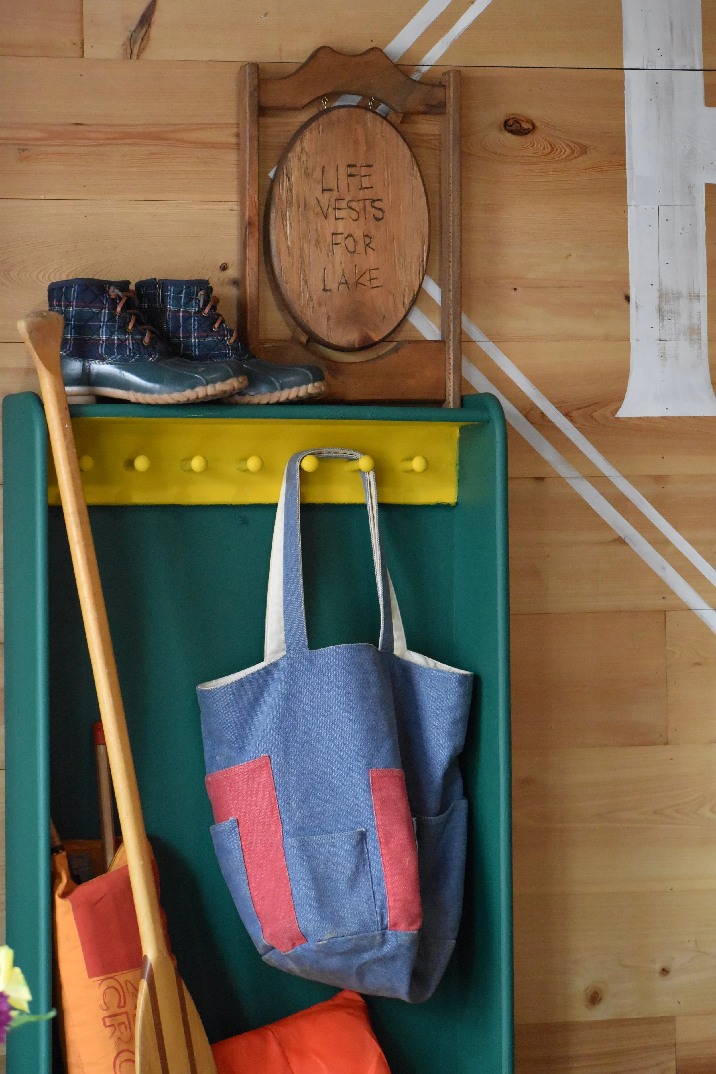 Wooden sign that says "LIFE VESTS FOR LAKE" on a wall with wooden paneling, above a yellow shelf, a blue bag hanging from a hook, a paddle leaning against the wall, a pair of plaid boots on a green surface, and parts of other items. Hamp & Harry's
