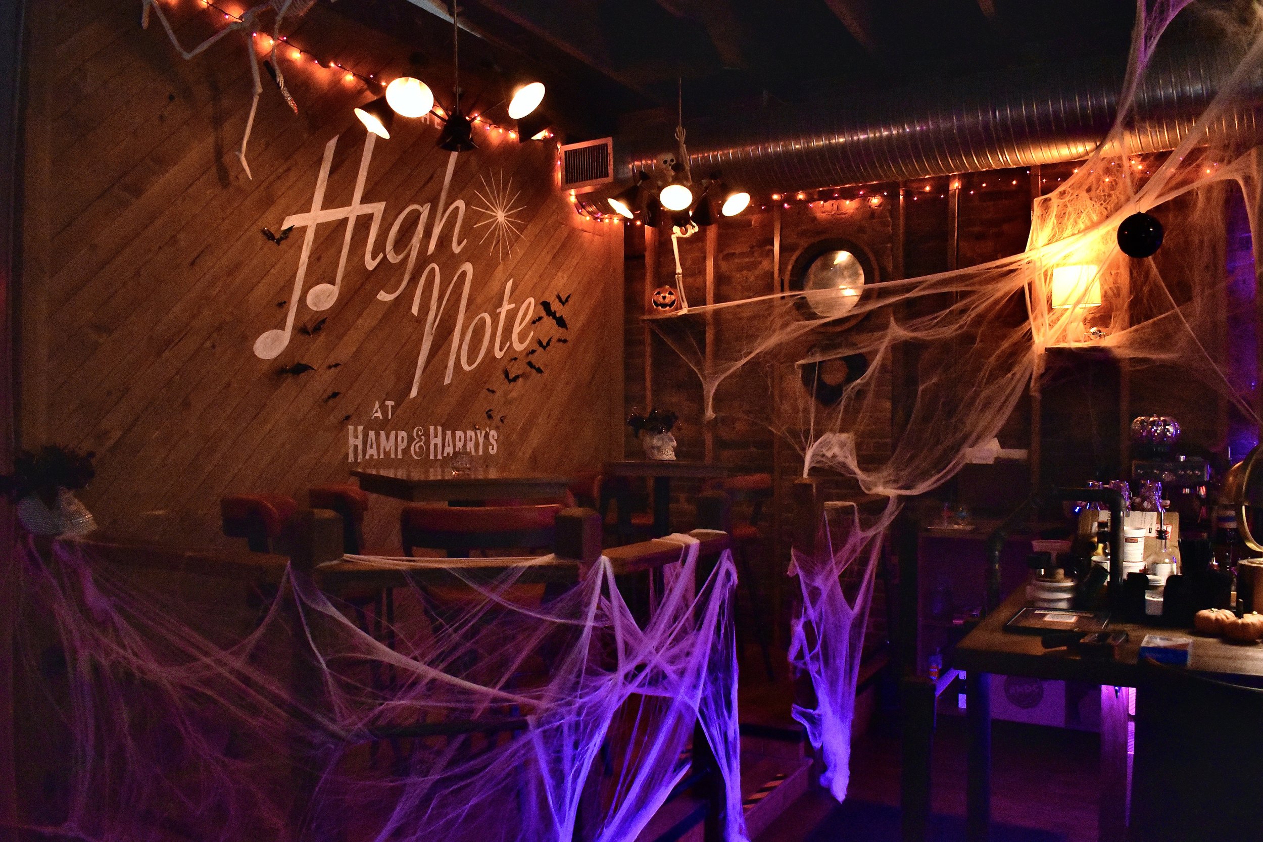 The interior of a bar or restaurant decorated for Halloween with cobwebs, spider decorations, orange and purple lighting, and a wooden wall with a sign that says 'High Note' and 'at Hamp & Harry's'.