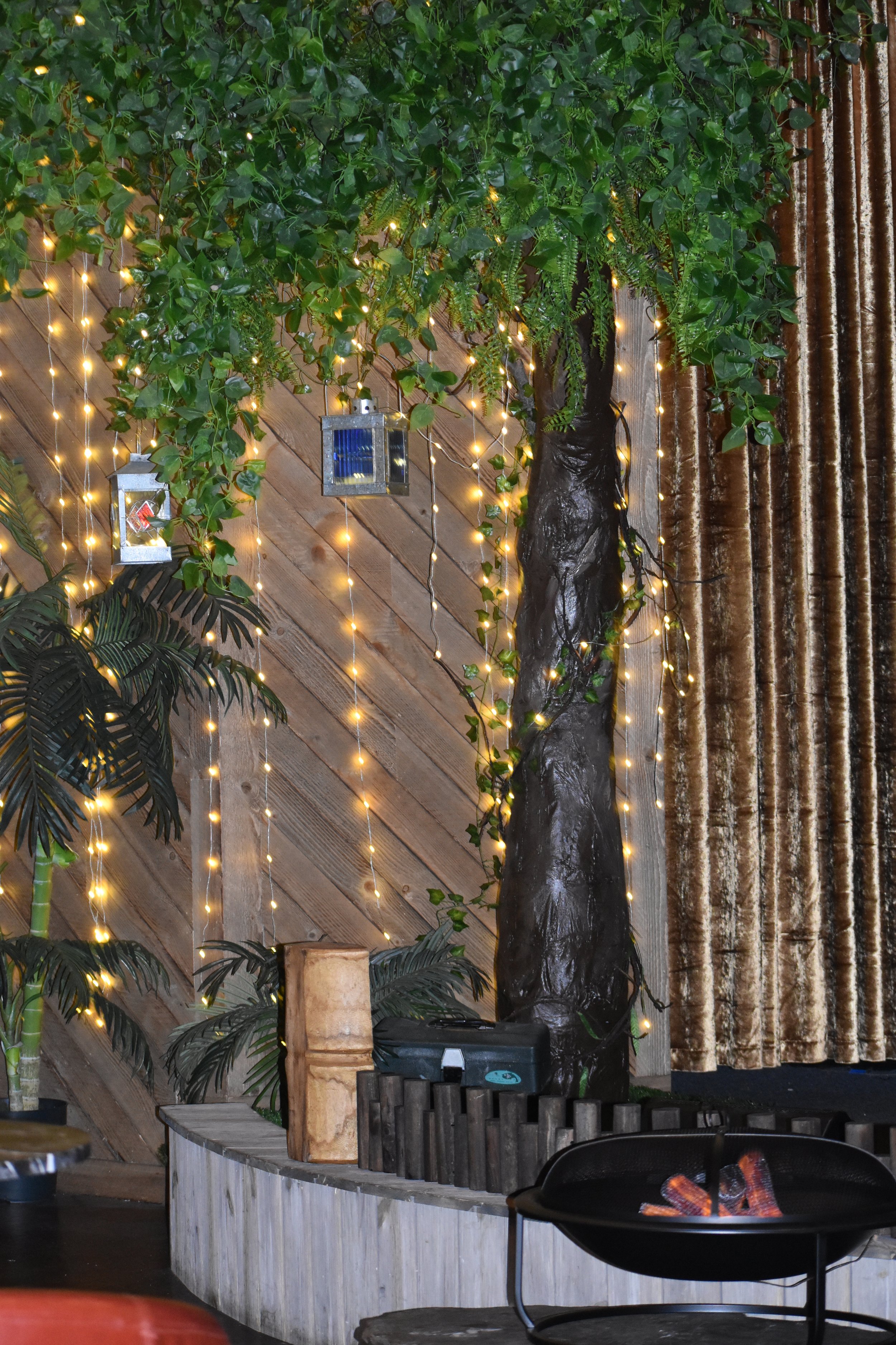 Indoor setting with artificial trees and plants, decorated with warm string lights and hanging lanterns, against a wooden wall and velvet curtains.