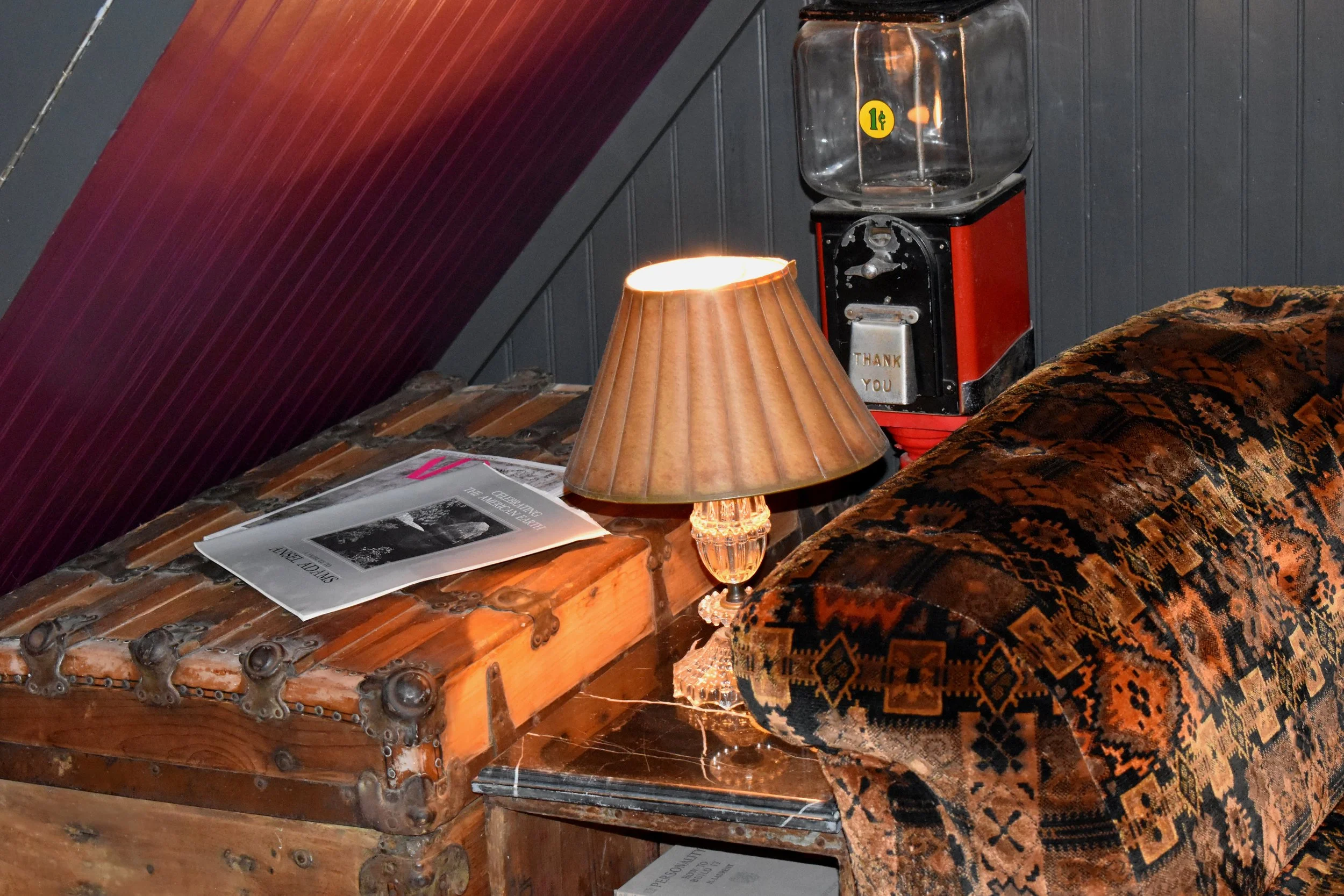 A vintage interior scene featuring a wooden chest with metal accents, an orange lampshade on a crystal base lamp, a vintage red vending machine with a glass container on top and a label saying 1¢, a patterned upholstered chair, and a small table with