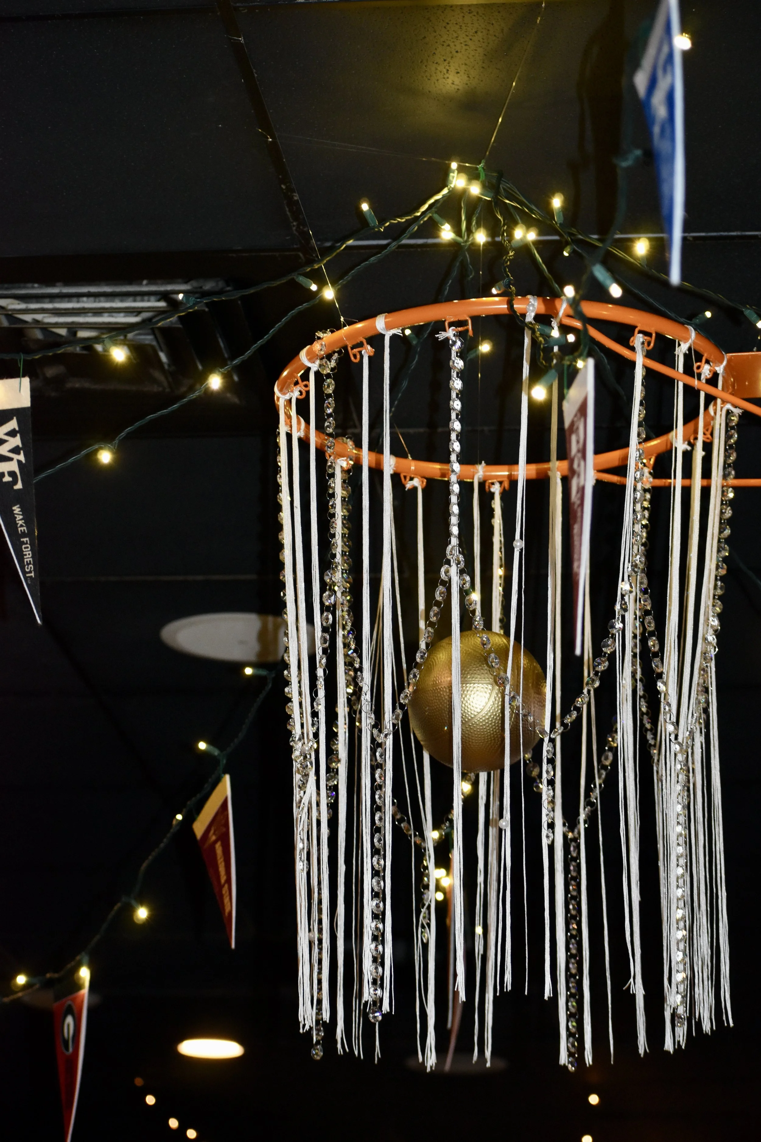 A decorative hanging ornament with white ribbons, rhinestones, and a large gold ball in the center, surrounded by small string lights, hanging from a ceiling. March Madness