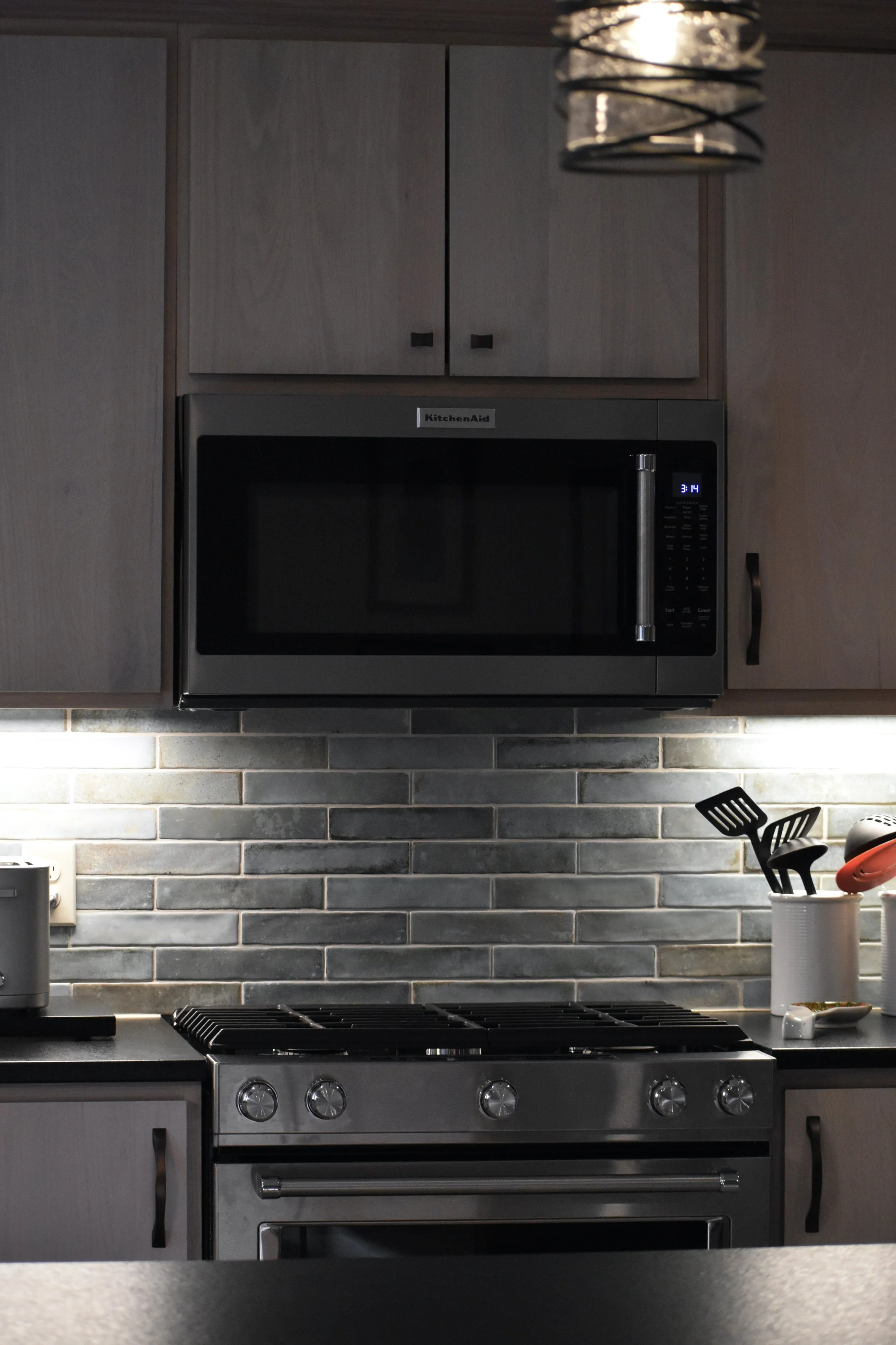 Modern kitchen with gray cabinets, black stove, and microwave. Under-cabinet lighting illuminates a gray tile backsplash. Kitchen utensils are in a container on the right, and part of a countertop is visible in the foreground. Marietta GA kitchen 