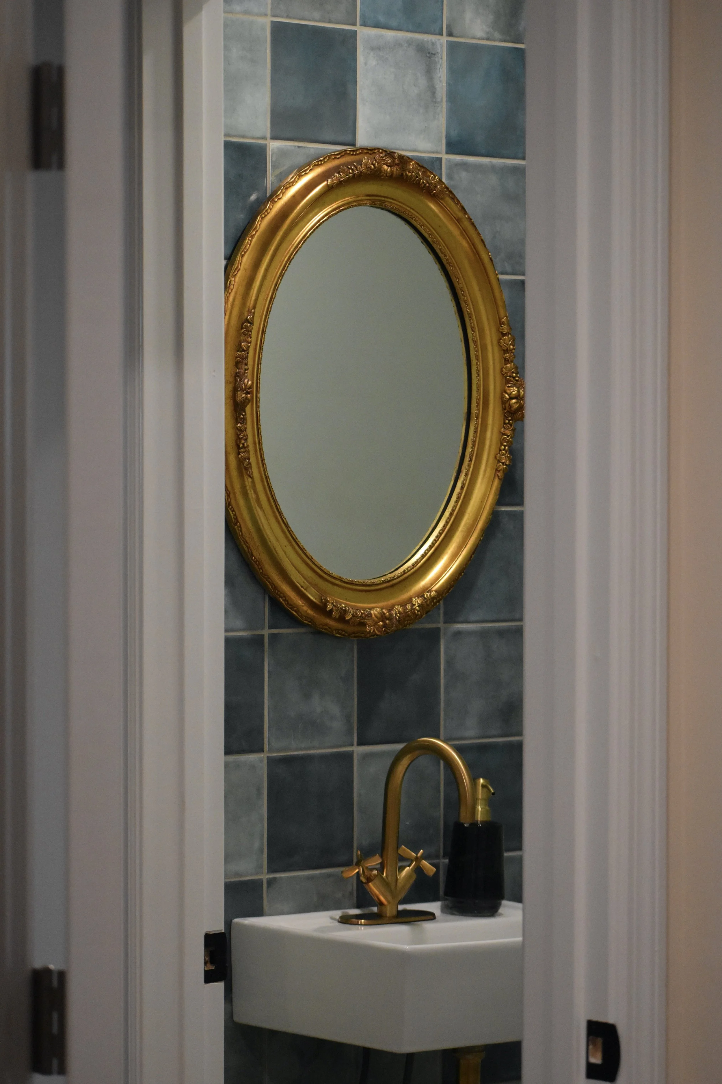 Bathroom with an ornate gold oval mirror, a small white sink, a gold faucet, and a black soap dispenser, with blue ceramic tile wall background. Marietta GA bathroom remodel.