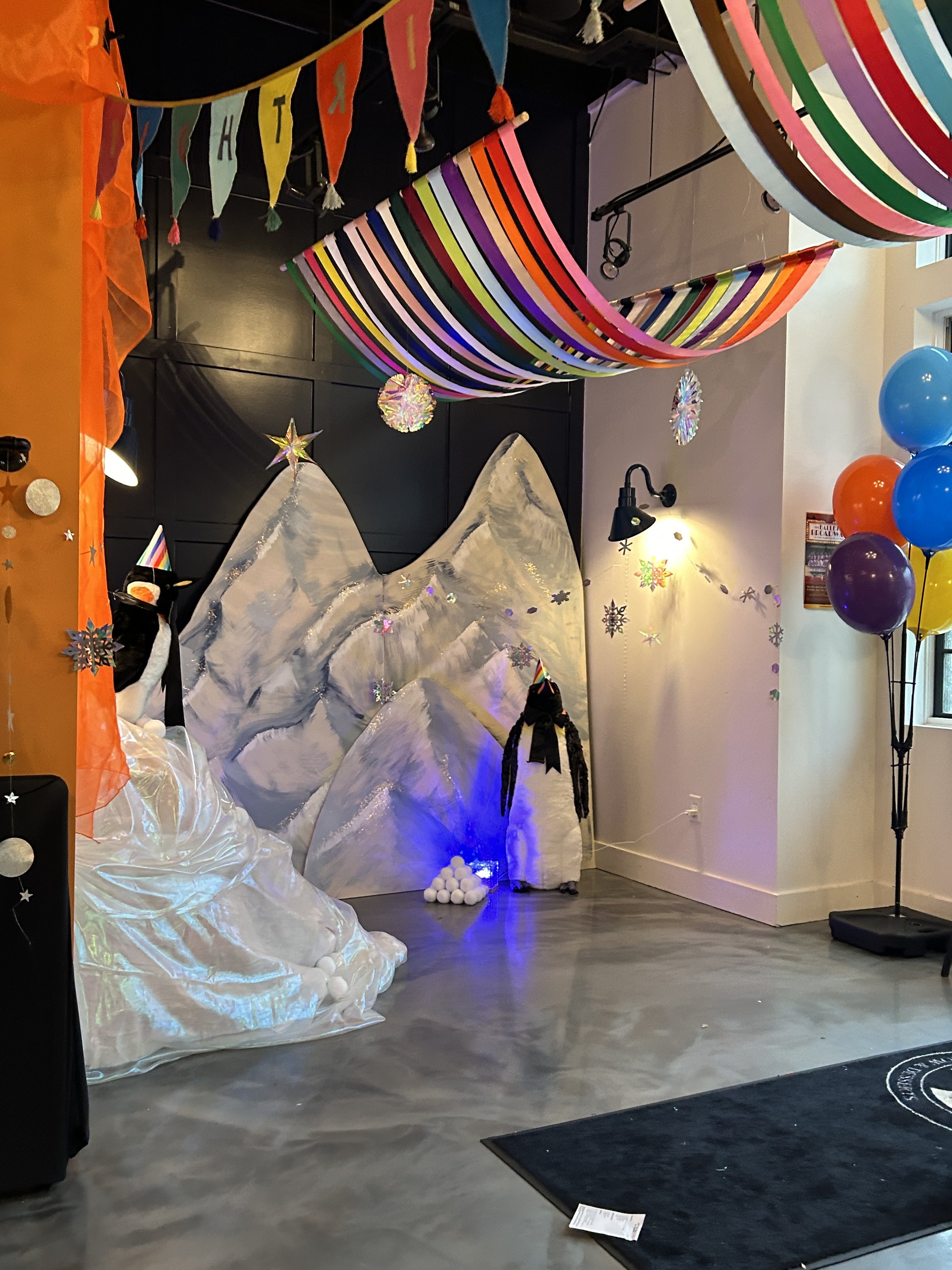Decorated indoor space with a mountain and penguin themed backdrop, colorful balloons, rainbow streamers, and festive banners for a celebration or party.