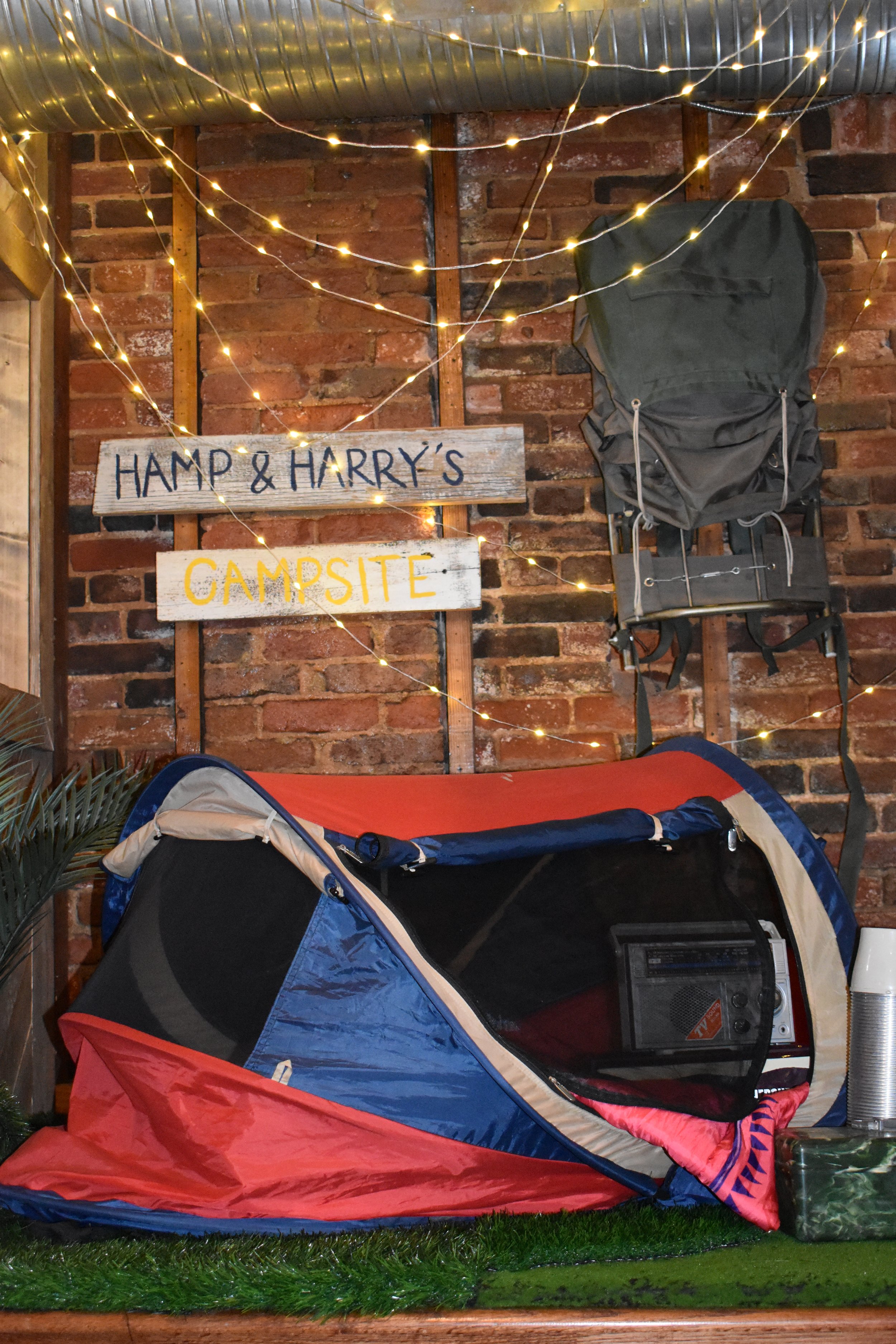 A camping setup with a small tent, a backpack stored on a wall, and string lights on a brick wall background. There is a sign that reads 'Hamp & Harry's Campsite'.