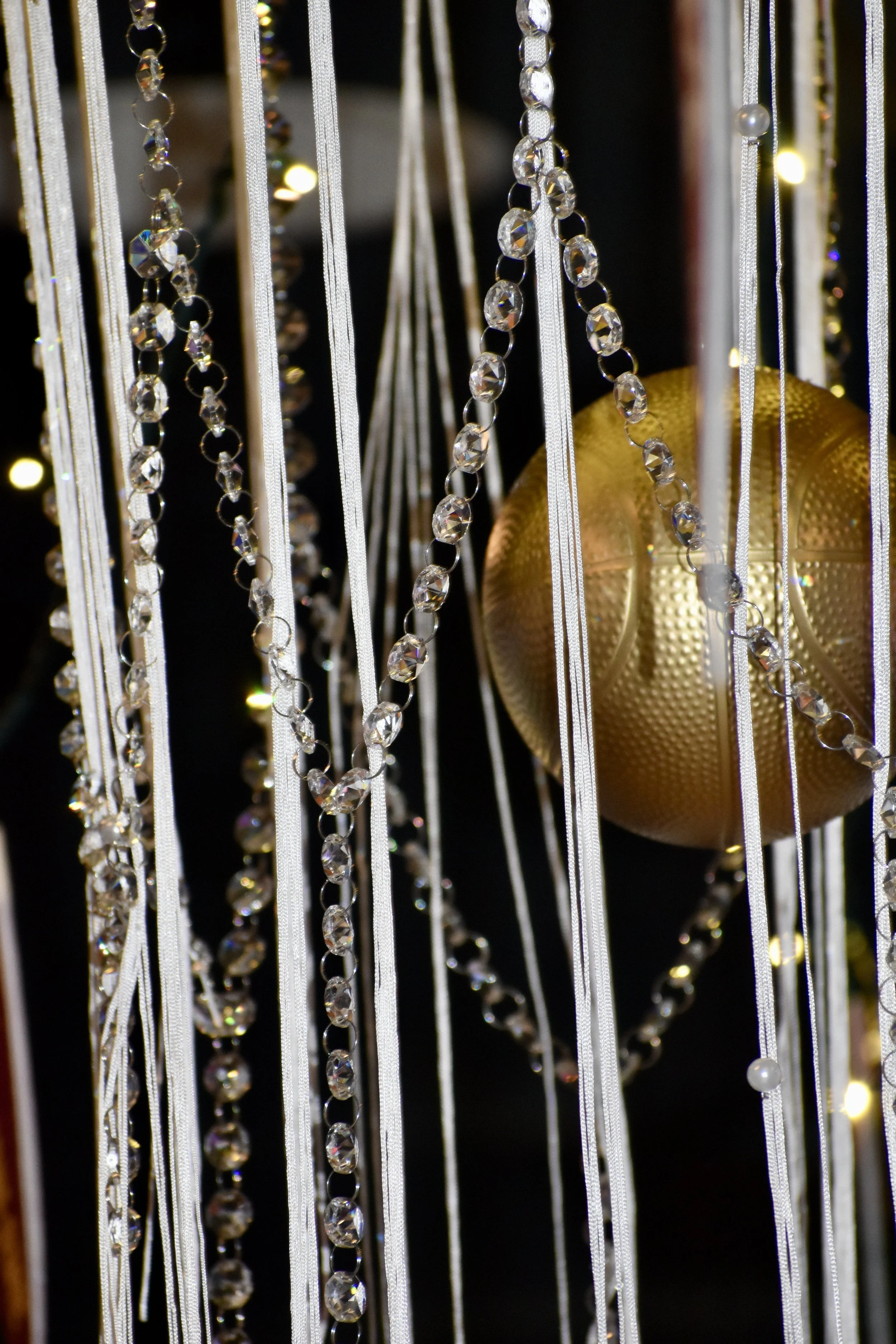 A close-up of a decorative Christmas ornament display featuring strings of clear beads, thin silver ribbons, and a large gold ornament with a textured surface. March Madness