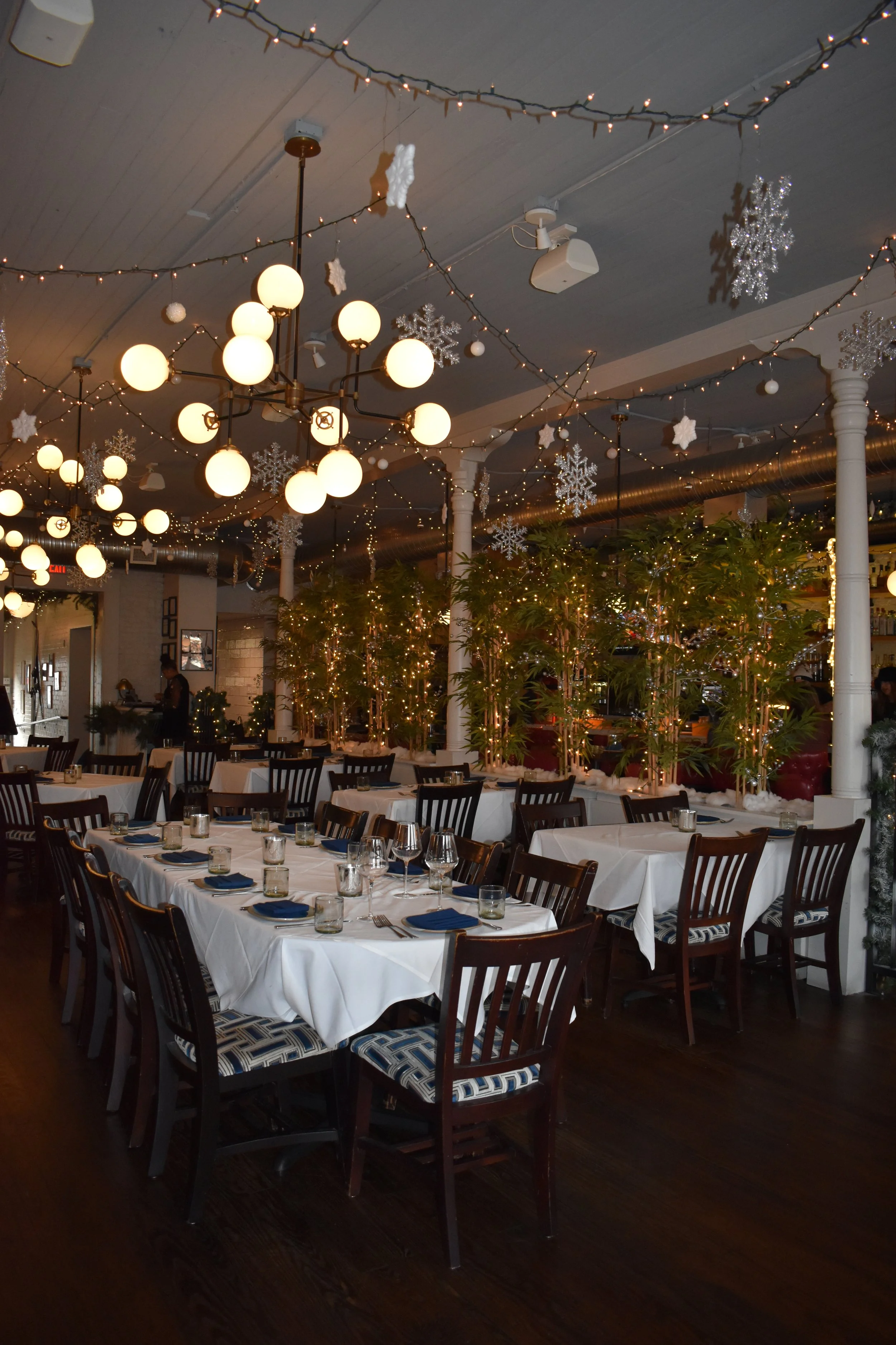 Festive restaurant interior with white tablecloths, blue napkins, glassware, and chairs, decorated with string lights, snowflakes, and holiday ornaments. Perry St Chophouse.