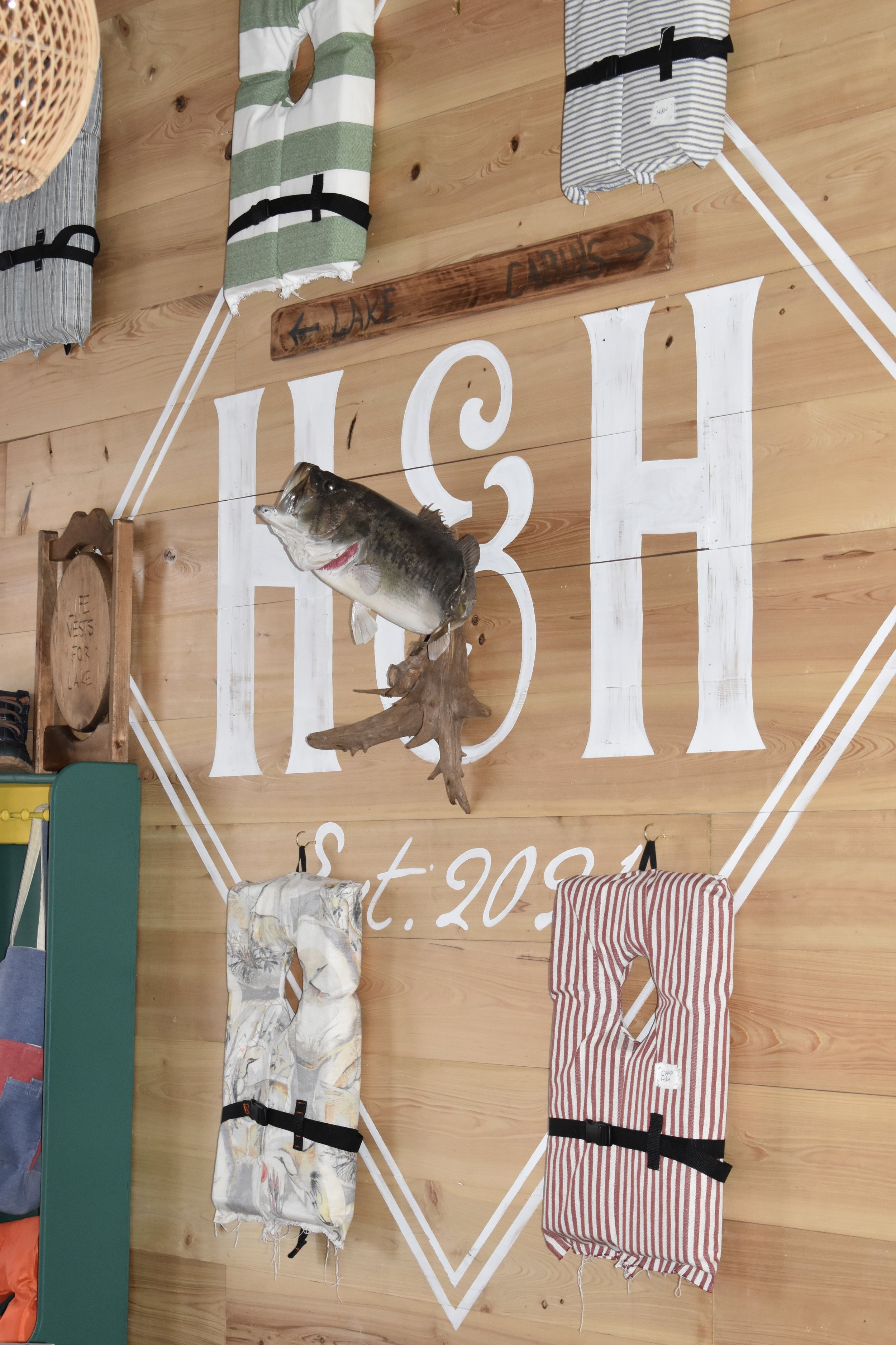 Interior of a store or restaurant with wooden walls and decorative fishing-themed items. There is a mounted fish hanging from the wall, a small wooden sign with directions to a lake and a public area, and colorful striped and fish-patterned fabric it