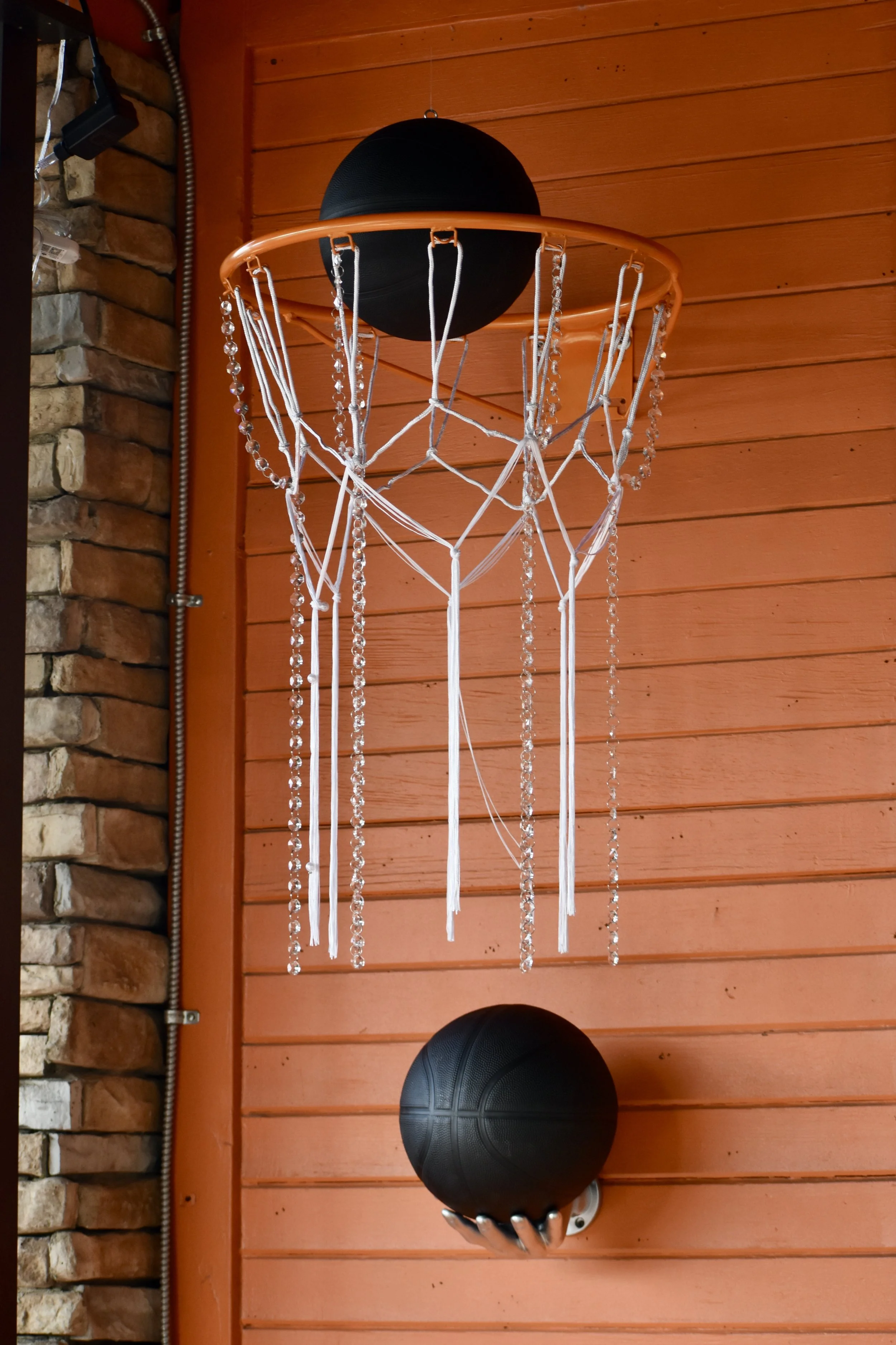A basketball hoop with hanging crystal decorations attached to the net is mounted on an orange wooden wall. A black basketball is held by a wall-mounted holder below the hoop. March Madness