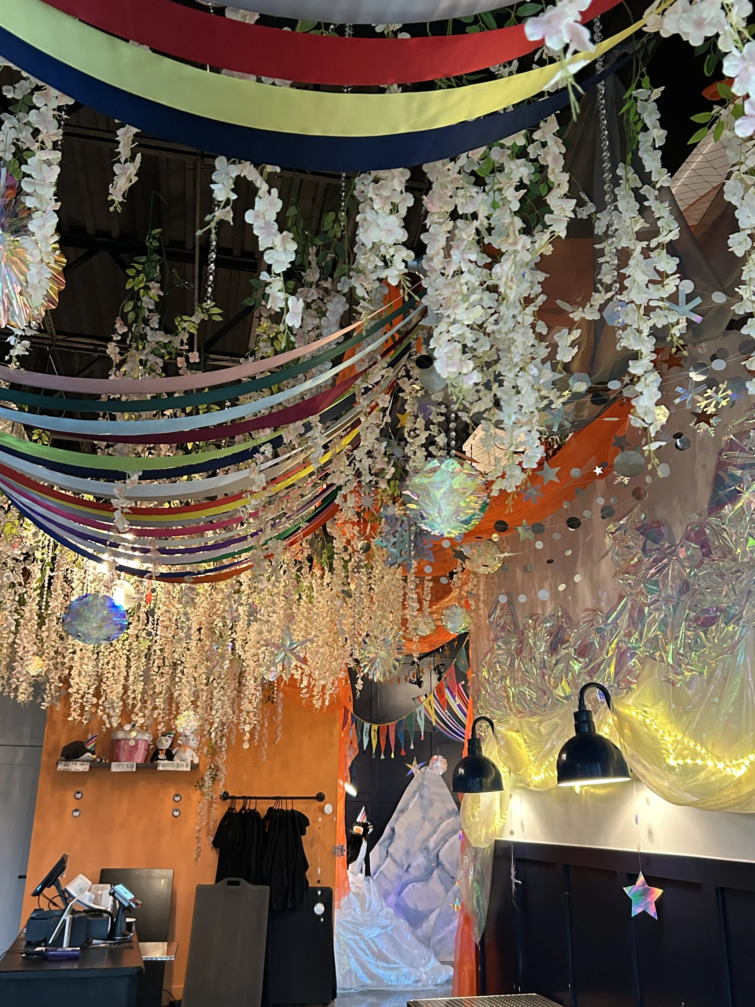 Decorative ceiling with colorful streamers, hanging white flowers, iridescent paper decorations, and star-shaped ornaments in a festive setting.