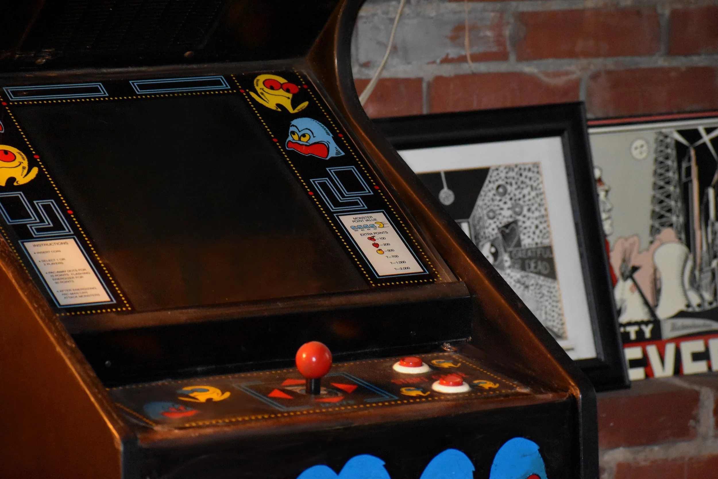 Close-up of a vintage Pac-Man arcade game machine with colorful graphics and controls, next to framed artwork on a brick wall
