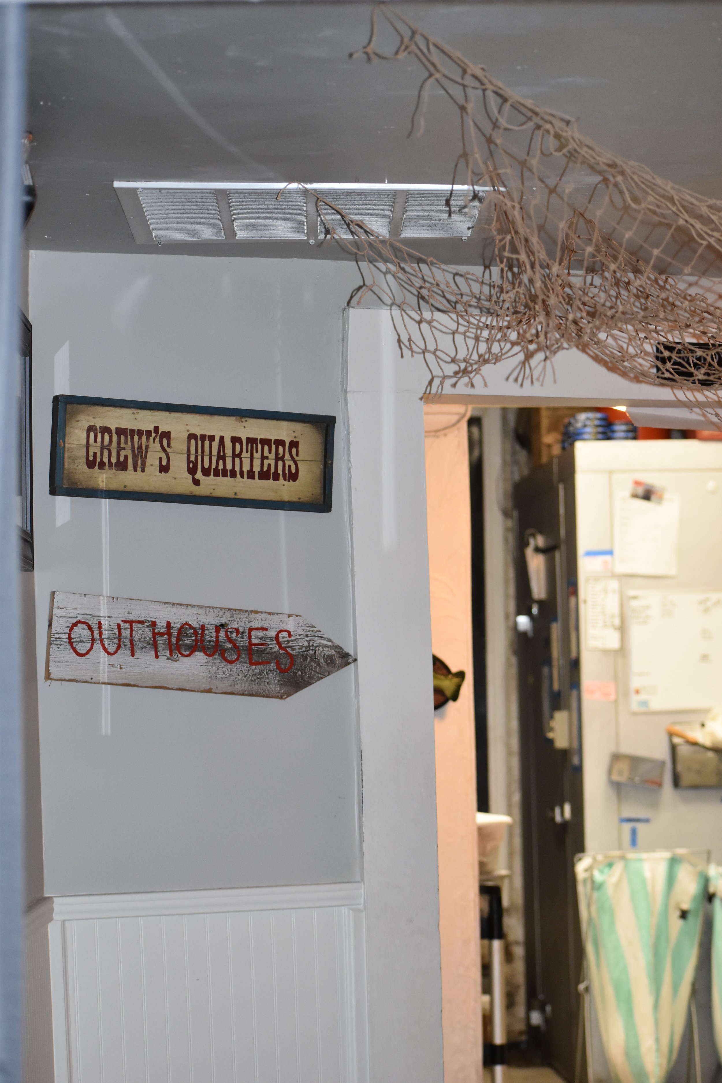 Interior view of a building with signs that read 'CREW'S QUARTERS' and 'OUTHOUSES' pointing to the right. There is a brown net hanging from the ceiling and part of a closet or storage area visible in the background. Hamp & Harry's Marietta Square.