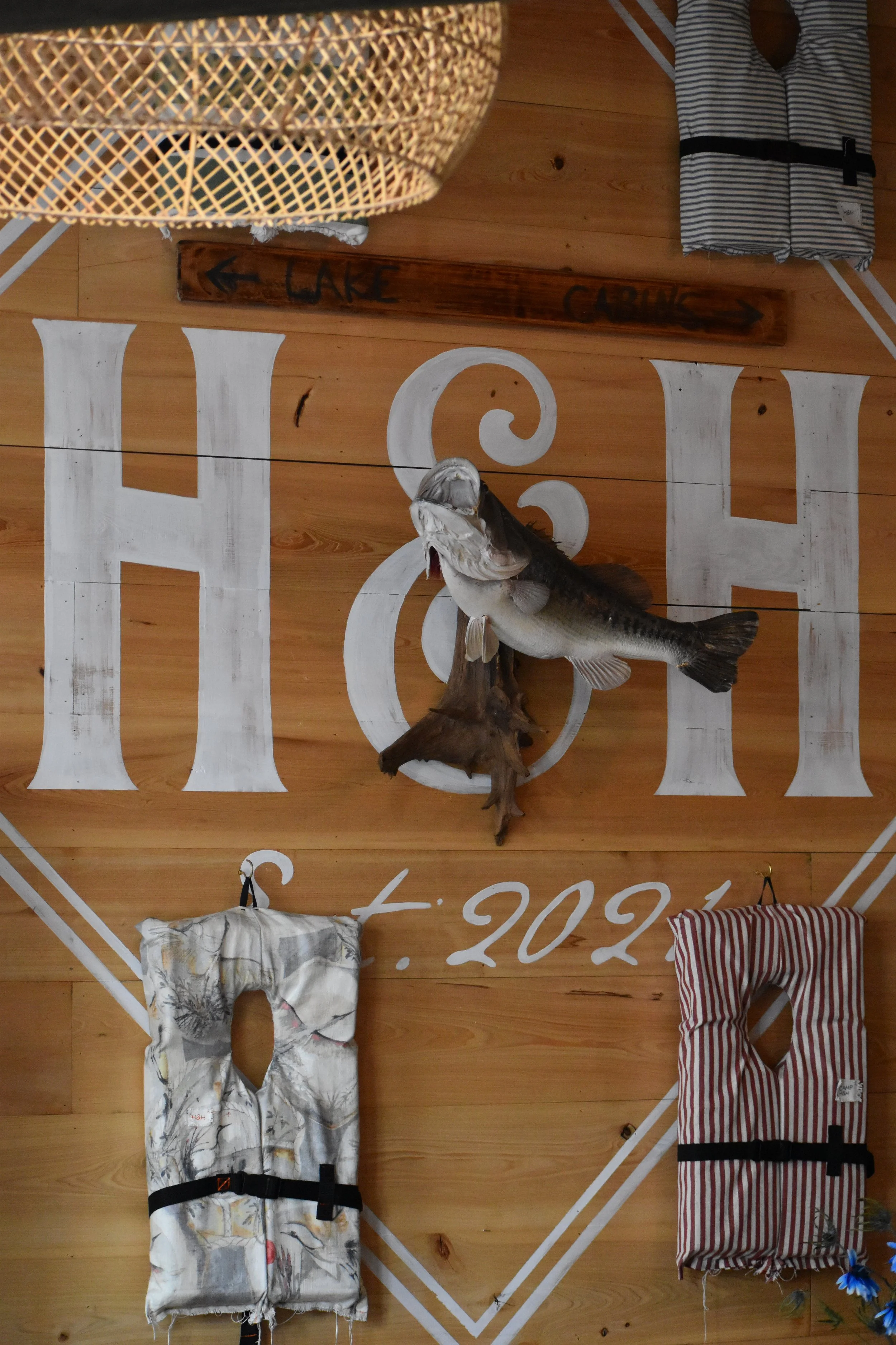 Decorative wooden wall with mounted fish and hanging fabric items, including a striped bag and a patterned cloth, with a corner of woven basket at the top and handwritten signs pointing to a lake and camp. Hamp & Harry's Marietta Square.