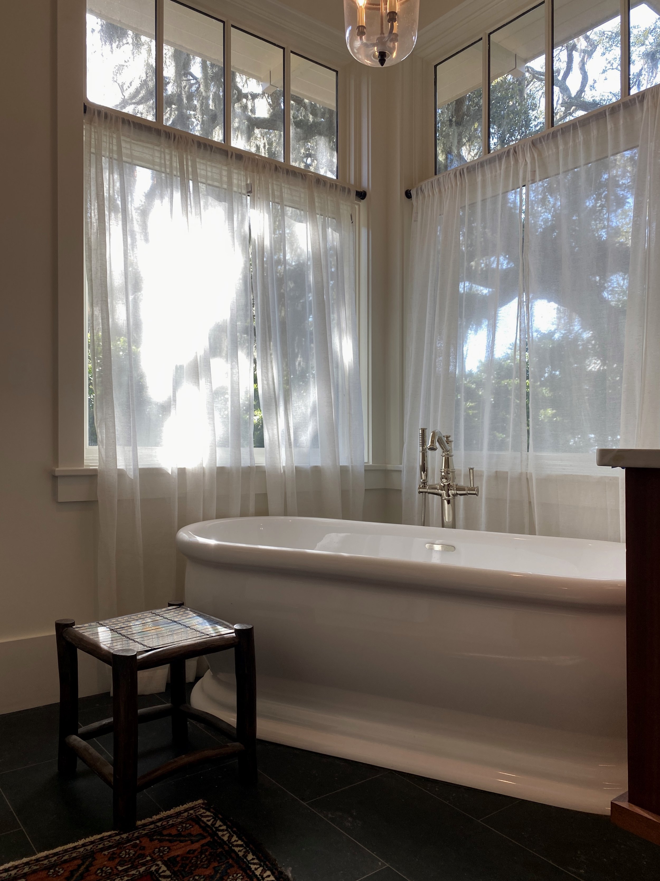 A cozy bathroom with a white freestanding bathtub beside large windows covered with semi-sheer curtains, a wooden stool, and a patterned rug on dark tile flooring. Upscale eclectic beach house bathroom suite.