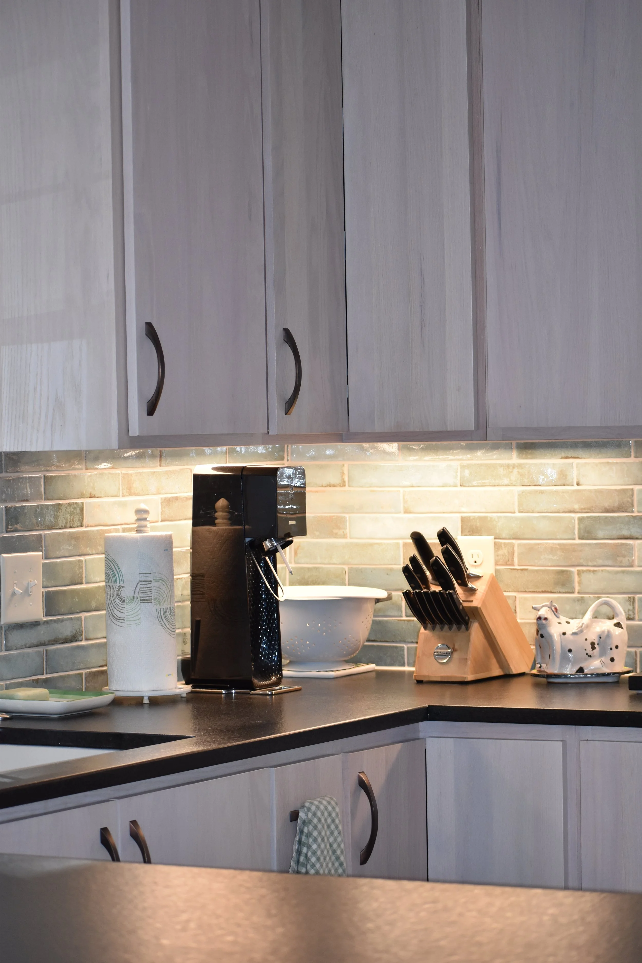 A kitchen countertop with a black coffee maker, a paper towel roll, a white bowl, a knife block, and decorative ceramic cow with black spots, with light wood cabinets above and a textured tile backsplash. Marietta GA kitchen remodel.